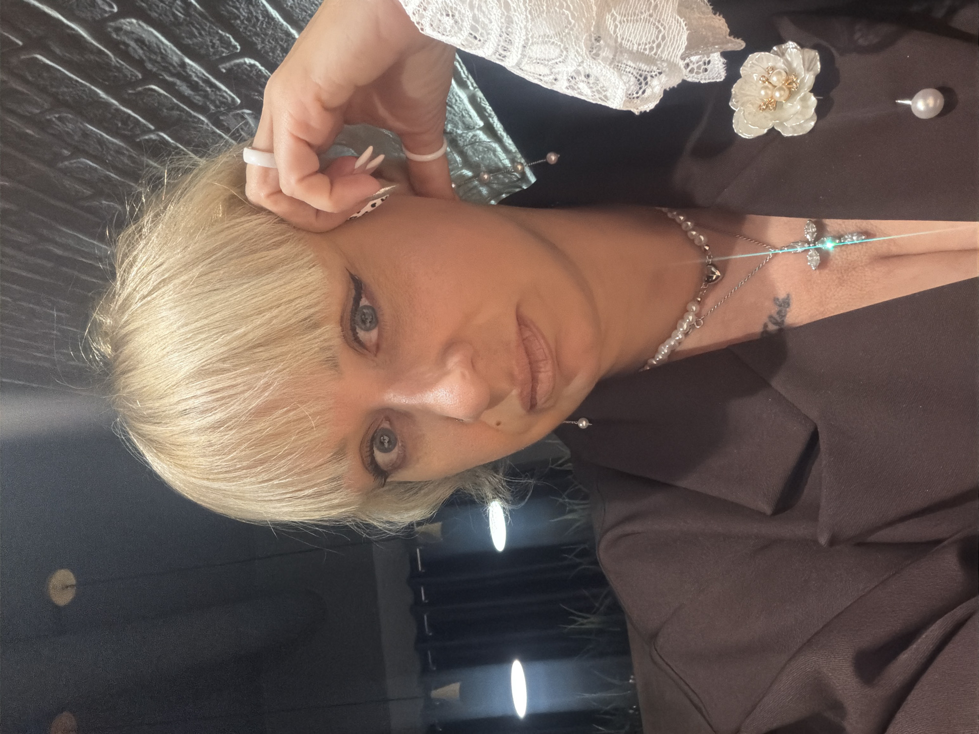 A close-up, portrait-style photograph features a woman with blonde, shoulder-length hair and bangs, looking directly at the camera. She has light-colored eyes and is wearing dark eyeliner and lip makeup. Her right hand, adorned with a white ring, is positioned near her ear, seemingly adjusting her hair or an unseen earring.
She is dressed in a dark-colored top or jacket, possibly brown or black, with a white lace trim visible on her shoulder or sleeve. Multiple pearl necklaces, including one with a cross-shaped pendant that has a sparkling effect, are visible around her neck and chest. A decorative white flower pin, also embellished with pearls, is attached to her clothing on the upper right side of the frame, next to a singular pearl stud or pin.
The background behind her head consists of a dark, textured wall, resembling brick or stone. Below that, the setting appears to be an indoor space with a dark, reflective surface showing bright, elongated light reflections, possibly from overhead lighting. The overall impression is that of a posed portrait in an interior setting, possibly in Chaikovsky, Russia. No visible text is present.