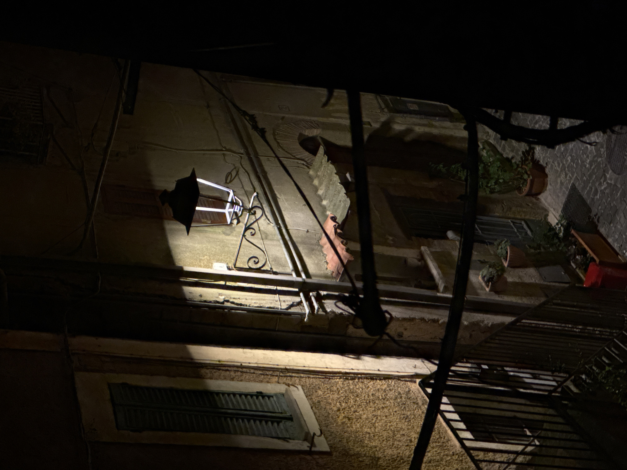 A nighttime shot, taken in Castelnuovo di Porto, Italy, of a building facade viewed from a low angle. The scene is predominantly dark, illuminated by an ornate, wall-mounted lamp with a white glow, casting light directly onto the building. Architectural features include windows with dark, louvered shutters, one directly below the light, and others higher up on the building. Electrical wires run along the walls. Potted plants sit on a windowsill. A metal structure is in the foreground. The street is cobbled.
