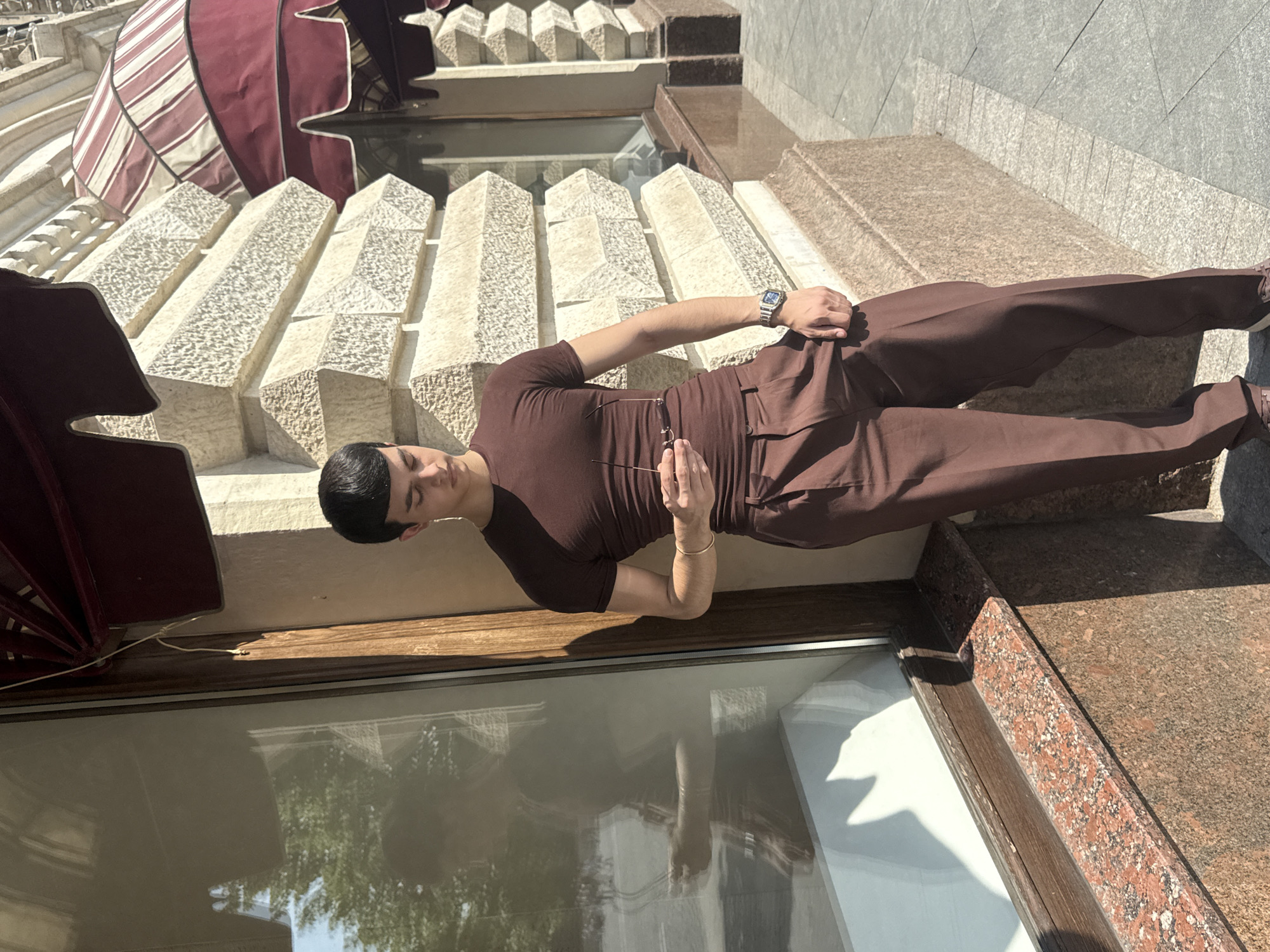 A man stands outdoors in front of a building in Unknown, Unknown. The man is wearing a brown outfit, consisting of a short-sleeved shirt and pants. He is holding a pair of glasses in his left hand and wears a watch on his left wrist. The background consists of a building facade with a canopy. The building's architecture features decorative columns and what appears to be a large window reflecting a tree. The ground and portions of the building are made of speckled brown stone.