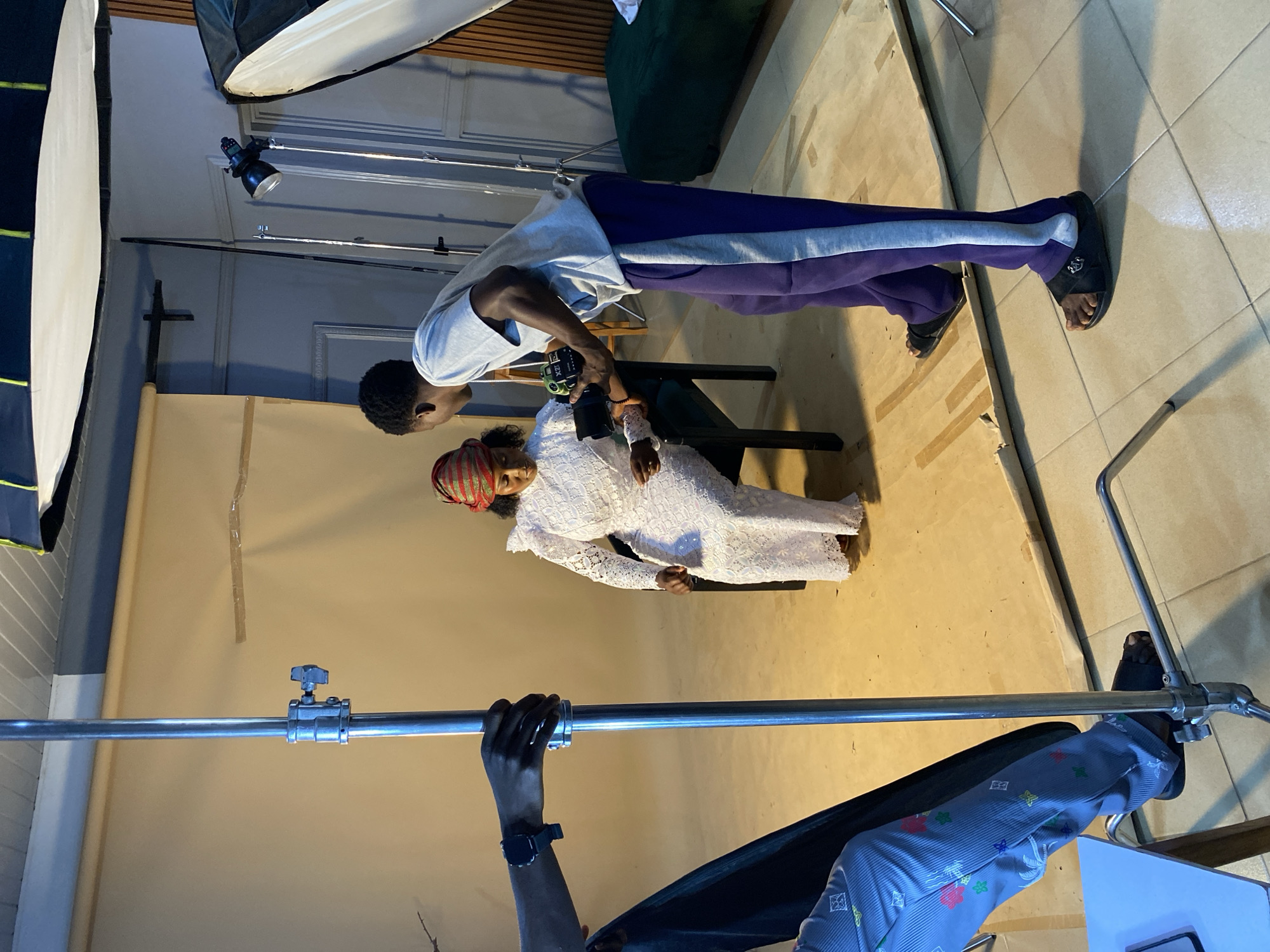 An indoor photoshoot is taking place in what appears to be a studio in Bukuru, Nigeria. The image, oriented sideways, shows two individuals and various photography equipment.
In the center, a male photographer, wearing a light grey t-shirt, purple sweatpants with a grey stripe, and black sandals, leans over a seated female model. He is holding a black DSLR camera with a large lens, seemingly adjusting it or reviewing a shot. The female model, seated on a dark chair, wears a white, intricately textured or embroidered dress and a red and green striped head covering. Her eyes appear closed or looking downwards.
The setting includes a light brown or tan backdrop, possibly a paper roll, and a tiled floor. Studio lighting equipment is visible: two large white softboxes are positioned at the top left and top right of the frame, indicating artificial illumination. A silver C-stand with a horizontal arm extends across the foreground at the bottom of the image, while another light stand is partially visible in the background. The walls of the room are light-colored. The overall scene suggests a professional and focused photographic session. No legible text is visible in the frame.