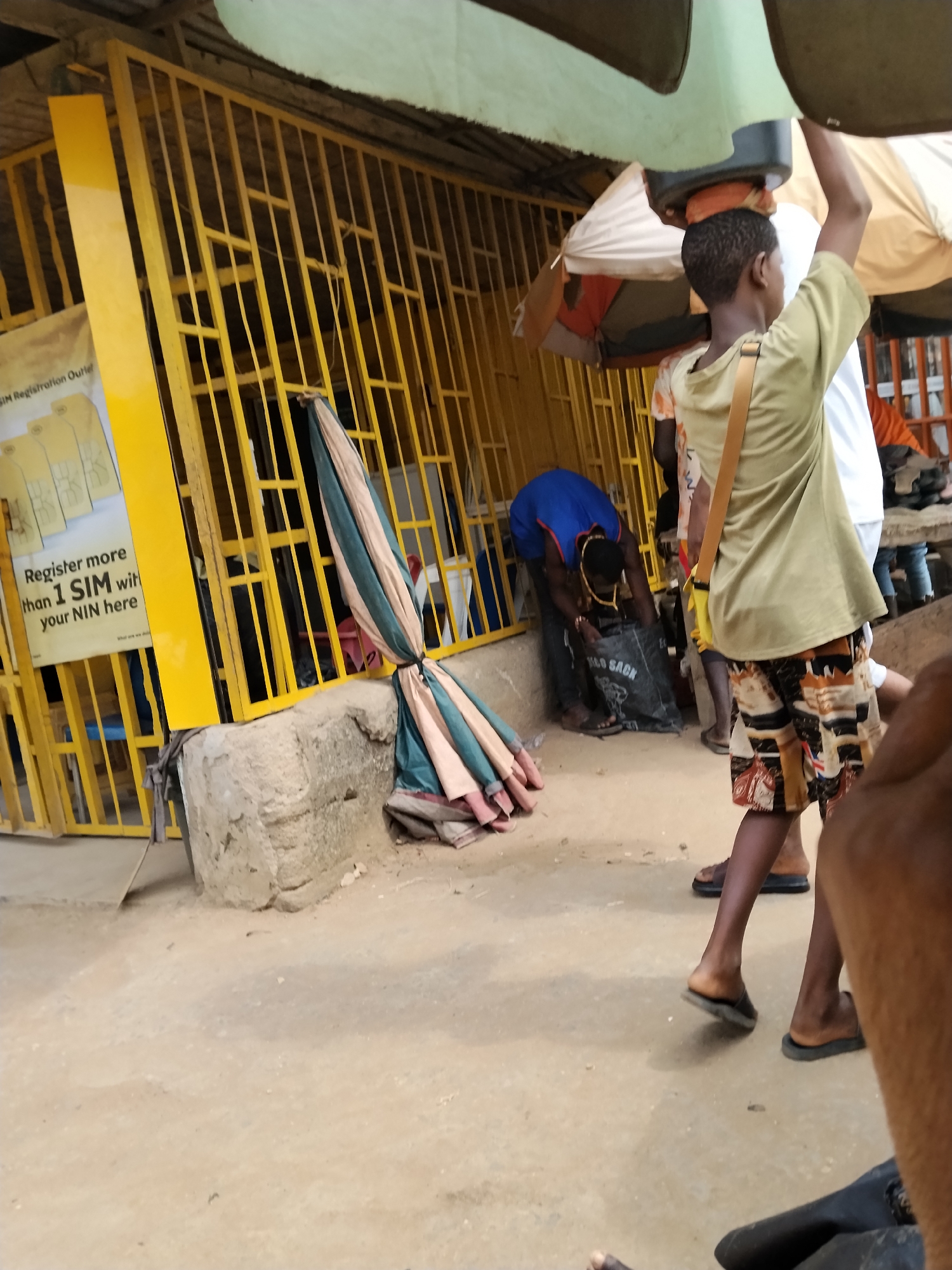 The image depicts a street scene in Ura Nta Umuarandu, Nigeria. A yellow metal structure, possibly a shopfront or entrance, dominates the left side of the frame. A sign is attached to it advertising SIM card registration. Several folded umbrellas are resting against the base of the structure.
A man in a blue shirt is bending over, appearing to rummage through a black "MOLO SACK." A young person with a bag strapped across their body is walking with a container balanced on their head. The background features other individuals, a partially visible white canopy, and a hint of further structures, suggesting a bustling environment. The ground is paved.