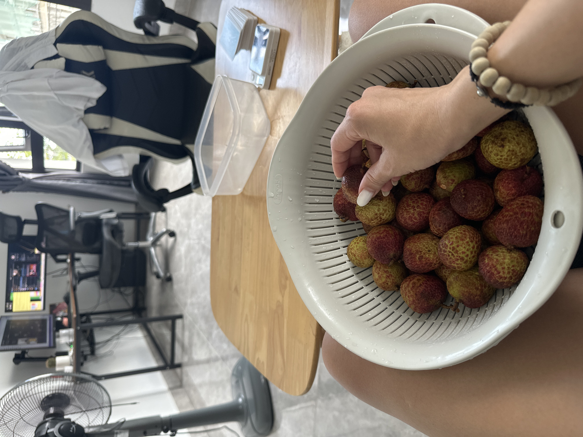 A person is holding a white colander full of lychees, picking one up with their right hand. The person is wearing a bracelet made of light brown beads. The colander is positioned over their legs. To the left, on a wooden surface, there is a clear plastic container and some smaller white objects. Behind this is an office setup with a desk, chair, and computer monitors. Further in the background, there's a black and white gaming chair with a white shirt draped over it. A grey fan is visible in the lower left corner. The location is a room in Đà Nẵng, Vietnam.