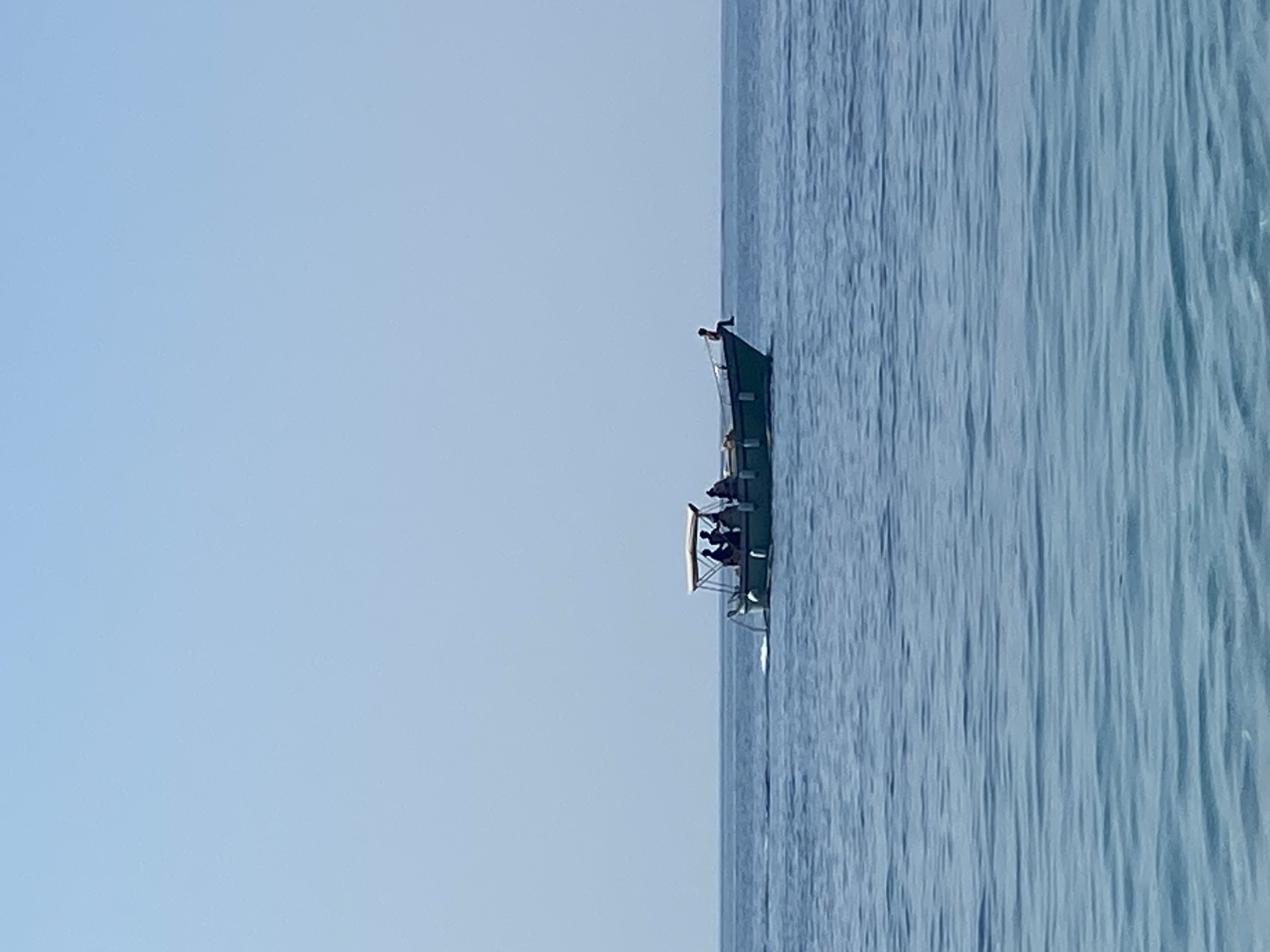 An eye-level, medium shot captures a small boat with a canopy sailing on a calm, blue sea. The boat, positioned slightly off-center to the right, has at least five people visible standing under the canopy, their silhouettes dark against the bright sky. The boat appears to be moving, with a wake trailing behind it. The horizon line is clearly defined, separating the vast, clear blue sky from the serene water. The overall environment is that of a bright, sunny day at sea. The location is identified as Prijevor, Montenegro.