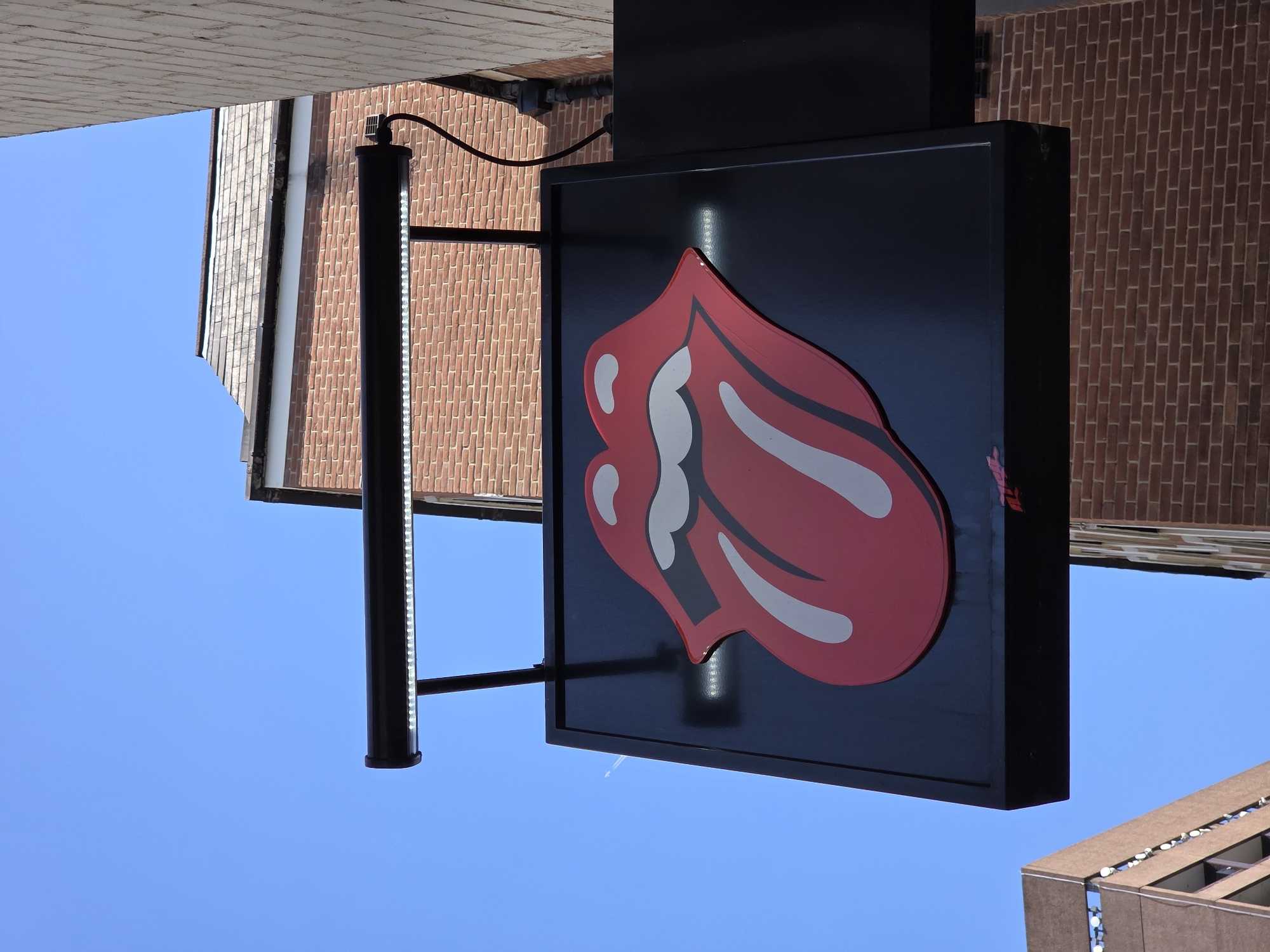 The image shows a Rolling Stones tongue and lips logo sign, illuminated and mounted on a building facade. The sign is predominantly black, with the iconic tongue and lips motif rendered in red with white accents. The sign is attached to the building with a black metal frame and a vertical light strip on the left side. The background includes sections of brick buildings and a clear blue sky. The buildings appear to be in an urban setting, potentially in London, United Kingdom, due to the specific style and placement of the sign, which aligns with the Rolling Stones' association with the city.