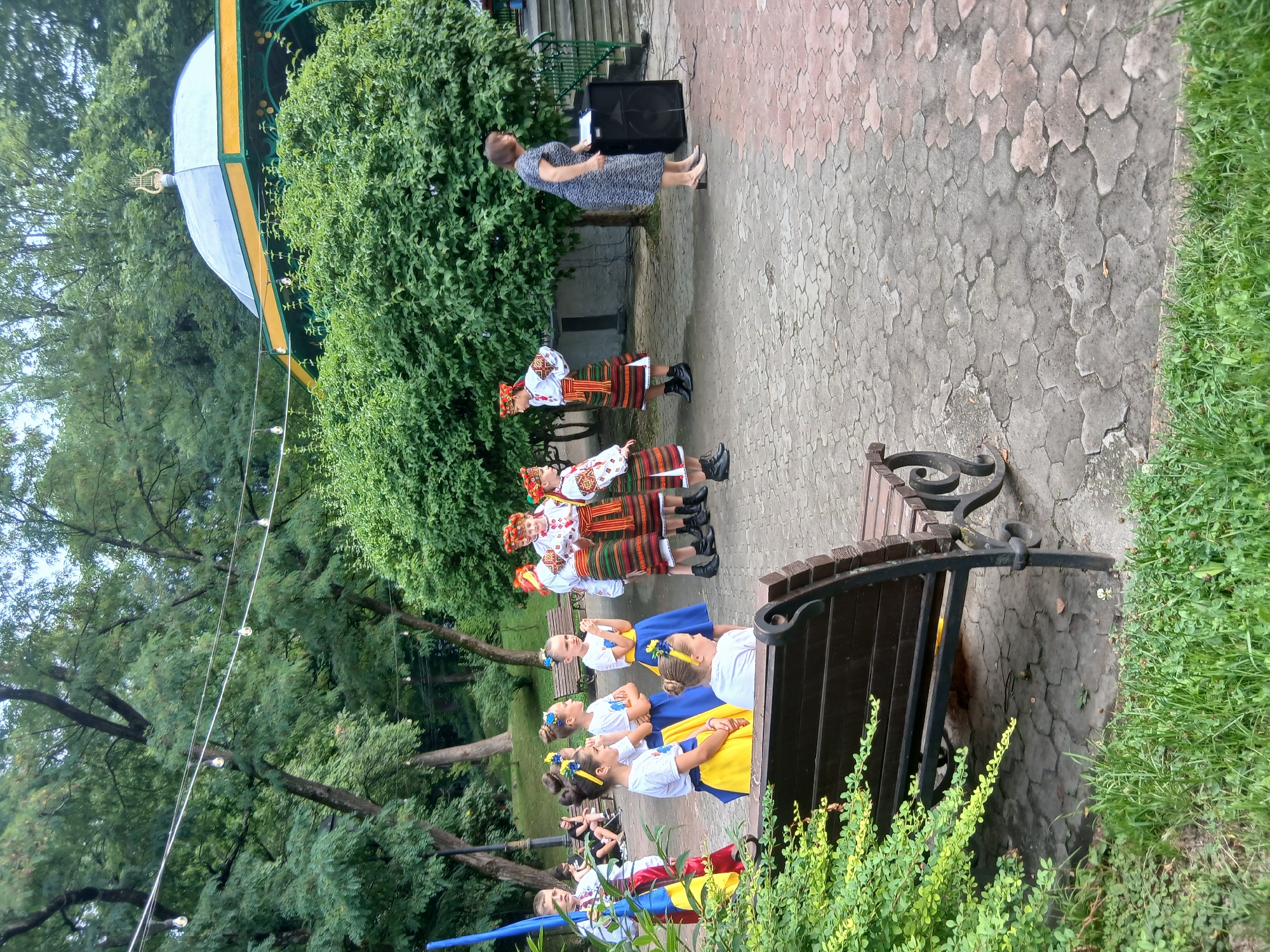 In Truskavets, Ukraine, a group of children in traditional Ukrainian attire, adorned with embroidered shirts and colorful skirts, stand in a park. Some wear flower crowns, and one boy holds a Ukrainian flag. They appear to be part of a performance or cultural event. To the right, a woman in a patterned dress stands near a large speaker, seemingly addressing the children or the audience. The setting is an outdoor paved area surrounded by lush green trees and bushes, with a park bench in the foreground. In the background, a decorative gazebo with a distinctive roof and a lyre-like ornament is visible, suggesting a public space designed for entertainment or gatherings.