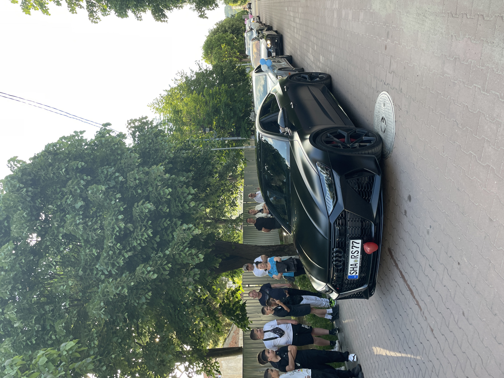 The image depicts a group of people gathered on a paved street next to a row of parked cars in Zlatograd, Bulgaria. The foreground features a black Audi RS model car parked alongside the curb, with its license plate visible, reading "SHA RS 77". A red heart-shaped balloon is attached to the front of the car. To the left of the vehicle, several individuals are standing, some of whom appear to be posing for a photograph. The group includes men, women, and children. The environment includes trees and foliage along the street, and the street is lined with other parked cars extending into the distance. A manhole cover is visible on the paved surface near the car.