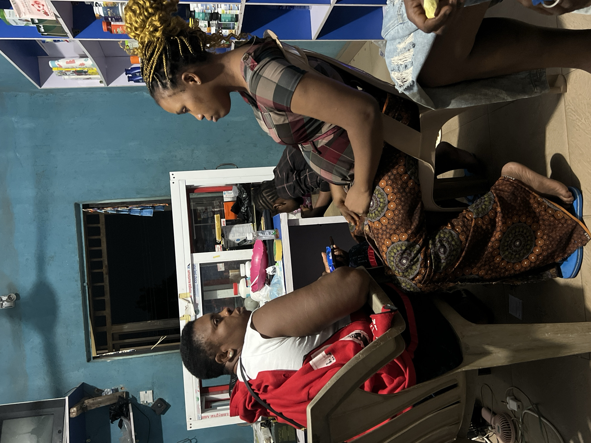 The image captures an indoor scene in what appears to be a local shop or business establishment in Obosi, Nigeria, likely during the evening or night due to the dark window and artificial lighting. The walls are painted a distinct blue-green.
At least four individuals are visible. In the foreground, a woman with braided, blonde-brown hair styled in a bun is prominent. She wears a plaid short-sleeved shirt (red, black, white, gray) over what looks like a dark, patterned overall skirt, and is leaning forward, looking down intently. To her right, partially visible, another person wears light blue ripped denim shorts and holds a small yellow object. Below the standing woman, the lower body of a person in a dark brown, circular-patterned (ankara) skirt and blue sandals is seen, sitting on a low stool or the floor. On the bottom left, a woman with short dark hair is seated on a light-colored plastic chair, wearing a white t-shirt with a red jacket draped over the chair. She looks towards the right, seemingly observing the activity. A fifth person in a dark plaid shirt is faintly visible in the background.
The setting includes blue and white shelves stocked with various products like bottles, boxes, and packaged goods, suggesting a retail environment, possibly a pharmacy or general store. A white-framed display cabinet or counter is on the right. A small window with metal bars is on the left wall, and a small fan and electrical outlet are visible on the floor.
The people appear to be engaged in a casual interaction or possibly a transaction. The standing woman's focused gaze suggests she is attending to something or someone directly in front of her.
Visible text on the white frame of the display cabinet includes "Kongtai Aluminium A.I", "Oluwa is my secret", and "Mmy Great Alumi", repeated. The overall ambiance is casual and busy.