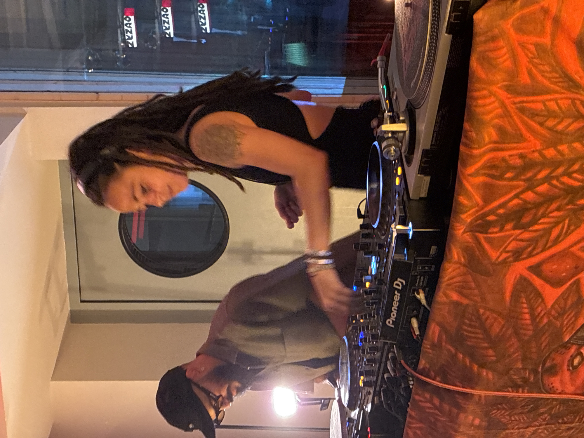 Two people, a man and a woman, are interacting with a Pioneer DJ setup with turntables. The woman has dreadlocks and a tattoo on her upper arm. The man wears a black cap and glasses. A patterned orange fabric drapes over the side of the DJ setup. Outside the window, vertical signs with the word "ZYZAO" are visible. The setting is likely a nightclub or bar in Barcelona, Spain, based on the "ZYZAO" signage which corresponds to a club in that city. The people appear to be DJing or involved in music performance.