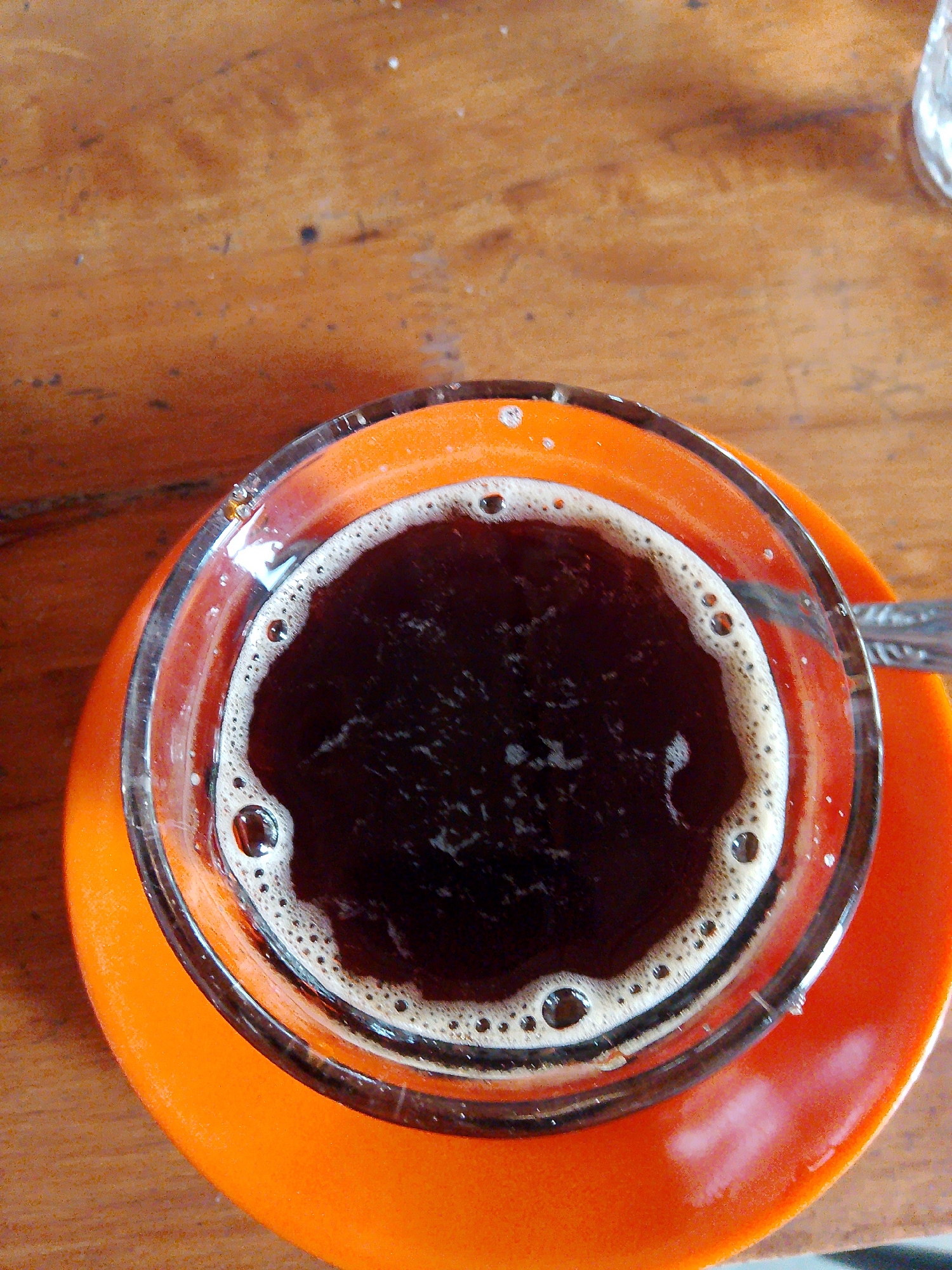 A top-down, medium shot captures a clear glass filled with dark coffee, sitting on an orange saucer. The coffee has a foamy, light brown rim with visible bubbles, and the dark liquid below shows some lighter, airy swirls. The glass and saucer are positioned on a wooden surface that appears to be a table, with a grainy texture and variations in color from light brown to dark orange. Part of a silver spoon is visible on the right side of the saucer, extending towards the cup. The lighting suggests an indoor setting, possibly a cafe or home. The image does not provide information about the specific location in Meunasahmee, Indonesia.