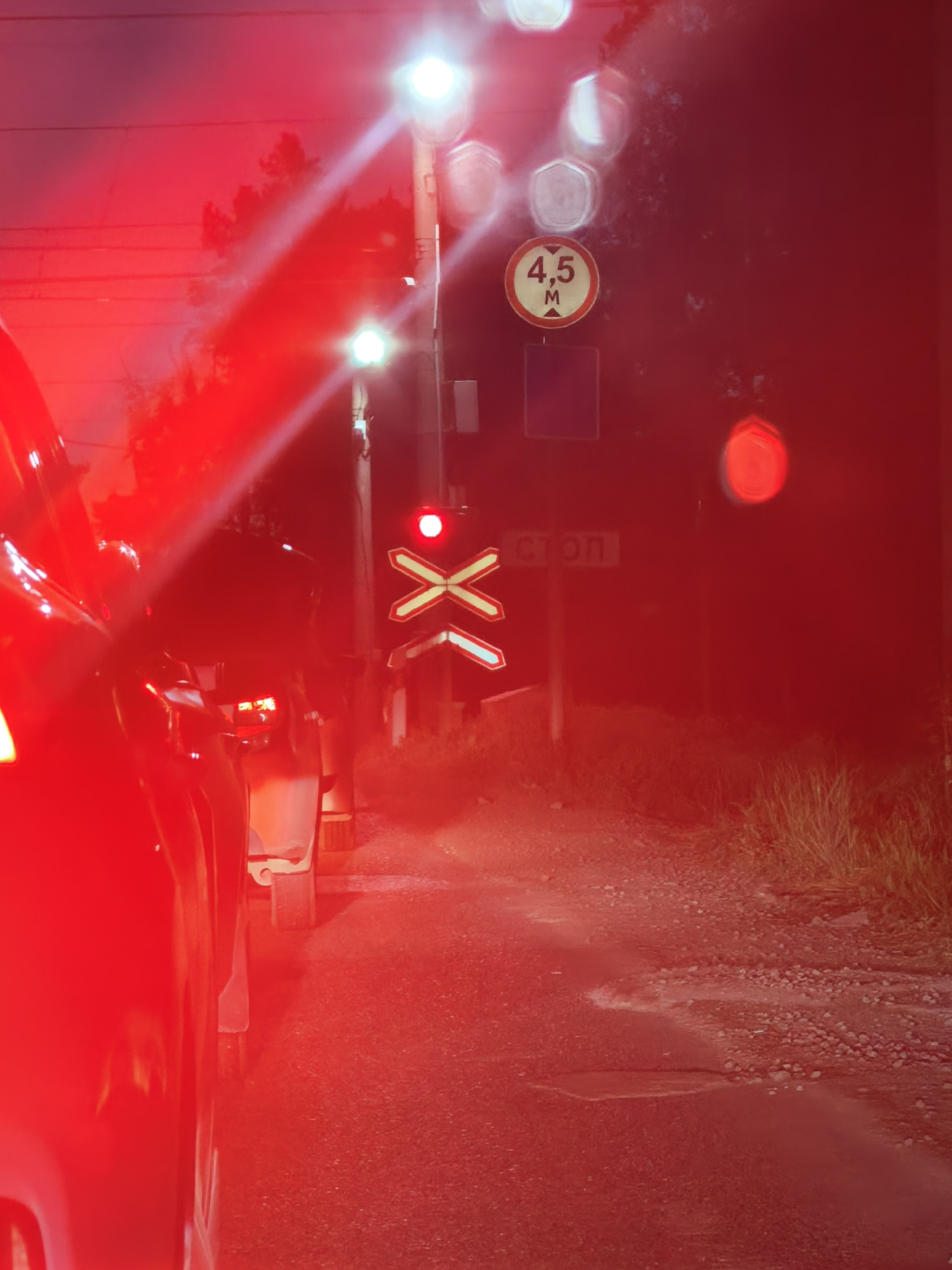 An outdoor nighttime photograph captures a red vehicle in the foreground, partially obstructing a railroad crossing. The crossing is indicated by a sign with white and red Xs. A red traffic light is illuminated above the crossing. To the left of the crossing, a circular traffic sign displays "4.5 M" within a white and red border. A smaller rectangular sign below it is not legible. In the distance, a sign with Cyrillic text "СТОП" is visible. The ground is a mixture of gravel and pavement, with some vegetation on the right side of the image. The overall scene is dominated by red light emanating from the vehicle and traffic signals, creating a hazy and somewhat blurred effect.