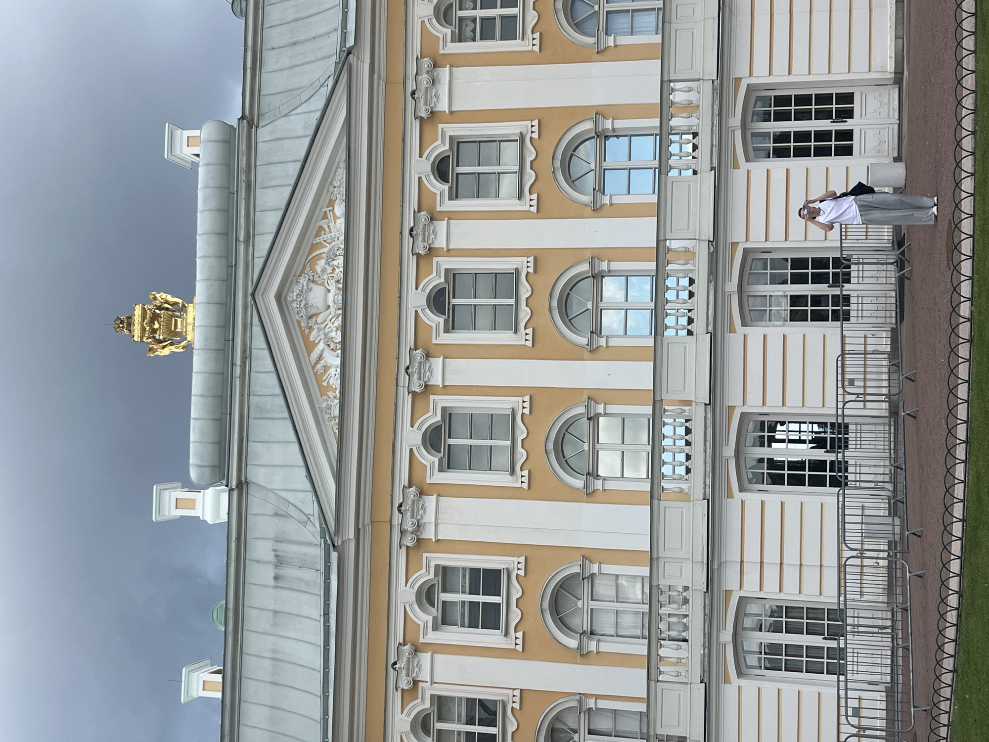A person stands in front of a large, ornate building with a yellow facade, white trim, and many windows. The building has a grey, pitched roof with decorative chimney-like structures and a golden crown-like ornament at the peak. The person, wearing a white t-shirt and grey pants, is holding up a camera and appears to be taking a photo of the building. There are metal barriers in the foreground, and a manicured lawn beyond them. The sky is overcast and grey. This is likely the Grand Palace in Peterhof, Russia, a historic imperial residence.