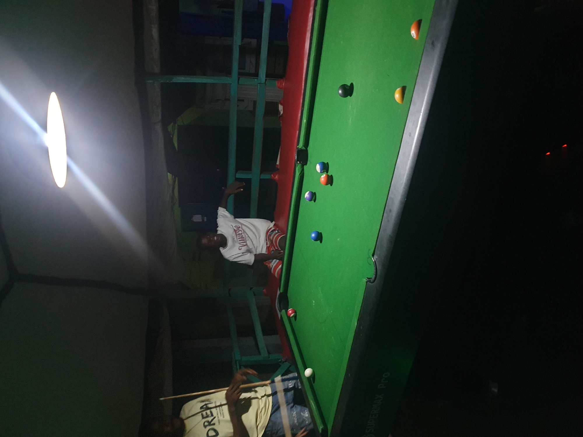 The image depicts an indoor billiards setting in Lokoja, Nigeria, likely a bar or recreational area. Two young men are present. One man, wearing a yellow t-shirt with "DREA" visible, stands on the left, holding a billiard cue. The other man, wearing a white t-shirt and patterned shorts, sits on a red bench to the right, facing the pool table. The pool table is green with several colored billiard balls scattered on its surface, including red, blue, orange, yellow, and a black ball. A bright overhead light illuminates the scene. The background shows a partially visible structure with a green railing. The overall atmosphere appears casual and relaxed, suggestive of a game in progress.