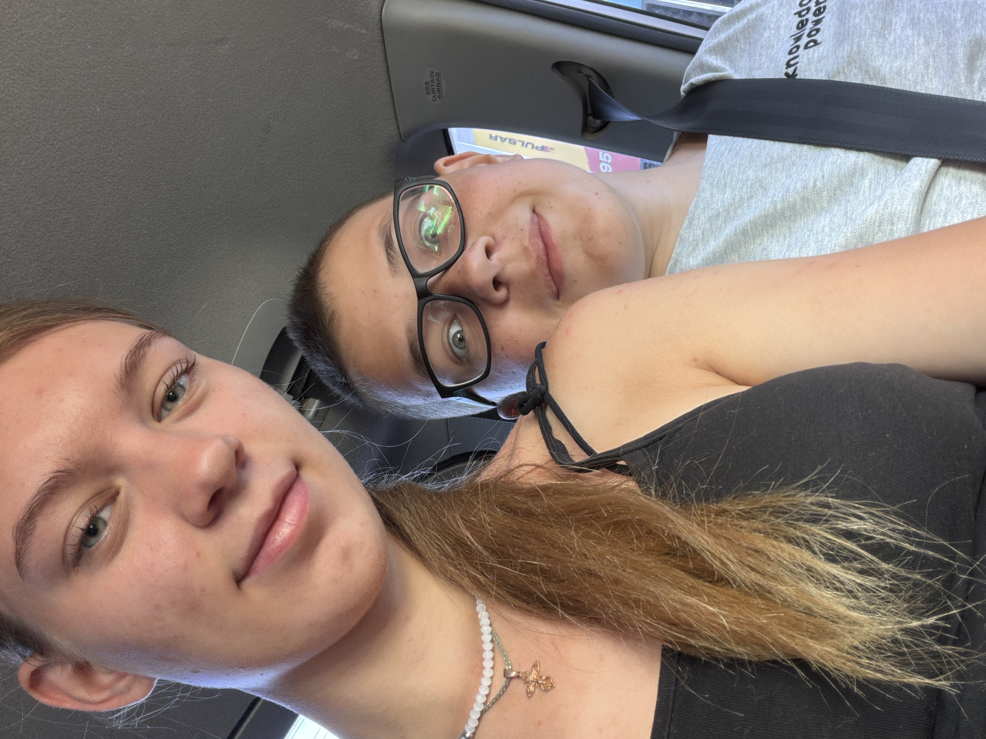 Two young people are in the front seats of a car. The person on the left, a female, is looking towards the camera. She has light skin, blonde hair pulled back, and is wearing a black tank top and a necklace with a cross pendant. The person on the right, a male, is looking towards the camera with a surprised expression. He has light skin, short dark hair, and is wearing glasses and a grey t-shirt. A black seatbelt is visible across his chest. The interior of the car is visible in the background. The environment appears to be a bright, possibly sunny day, given the reflections in the male's glasses. The image does not provide any specific visual cues to determine the exact location within Rostov-on-Don, Russia.