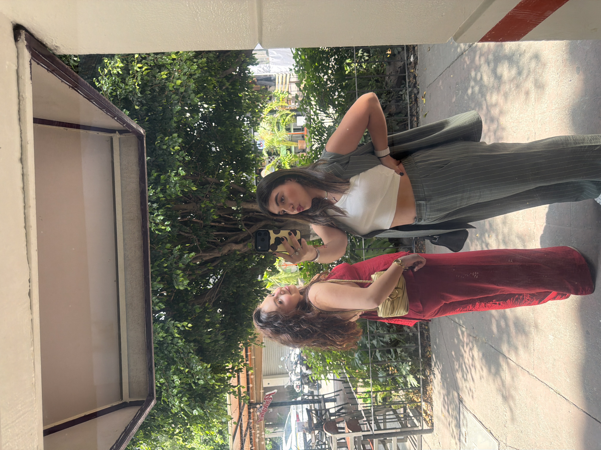 Two young women are posing for a selfie in front of a reflective surface. The woman on the right is wearing a white crop top, olive green pinstripe pants and a matching blazer. The woman on the left is wearing a long red dress with a beige shoulder bag. The reflection shows a street scene with trees, buildings, and outdoor seating. The presence of a "Kermesse" sign in the reflection, along with the architectural style of the buildings, suggests the location is in Mexico City, Mexico.