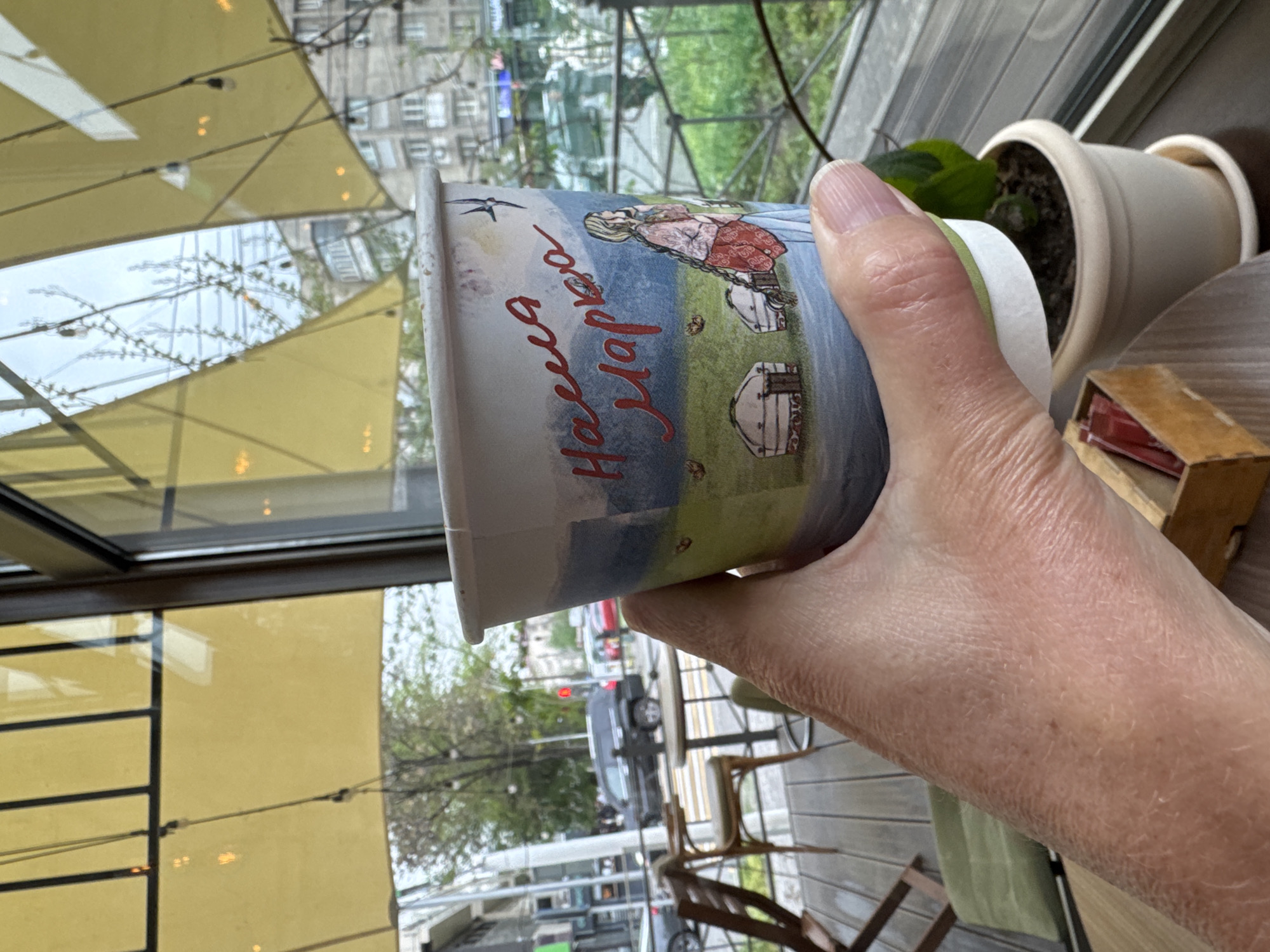 The image, rotated 90 degrees counter-clockwise, captures a person's hand holding a disposable cup while looking out from an indoor setting onto a city street. The hand firmly grips the cup, which is decorated with an illustration of a woman in traditional dress, several yurts, and a bird, set against a blue and green landscape. Red Cyrillic text "Наша Марка" (Nasha Marka), meaning "Our Brand," is prominently visible on the cup.
Through the large window, reflections of yellow awnings or internal structures are visible. Beyond the reflections, an outdoor city scene unfolds, likely in Bishkek City, Kyrgyzstan. The view includes a street with a car and a red traffic light, and a multi-story building in the background. Trees with sparse, budding leaves suggest springtime. Adjacent to the building, an outdoor seating area with chairs and a table is visible.
Indoors, to the right of the cup, a small green plant in a white pot and a small wooden box, possibly holding sugar packets or matches, are placed on a wooden surface. The setting suggests a cafe or restaurant, where someone is enjoying a drink while observing the activity outside on a seemingly overcast day.