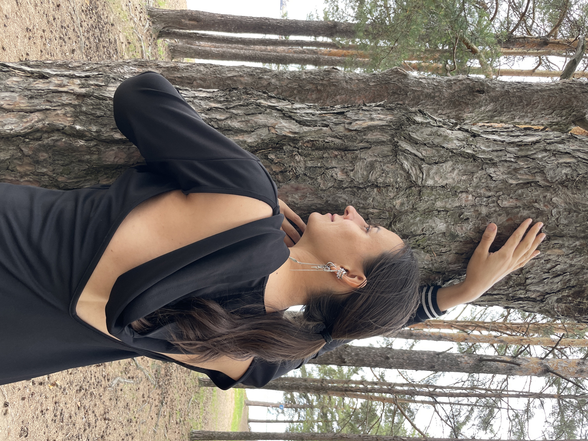 A woman stands in profile, her left arm resting against the trunk of a large pine tree, in a forest setting. The woman has dark hair pulled back in a ponytail and is wearing a black dress with a deep, open back and long sleeves. She is also wearing dangling silver earrings. The background consists of more pine trees, a body of water visible in the distance, and a forest floor covered in pine needles and cones. The sky is overcast. This scene is located in Chaikovsky, Russia.