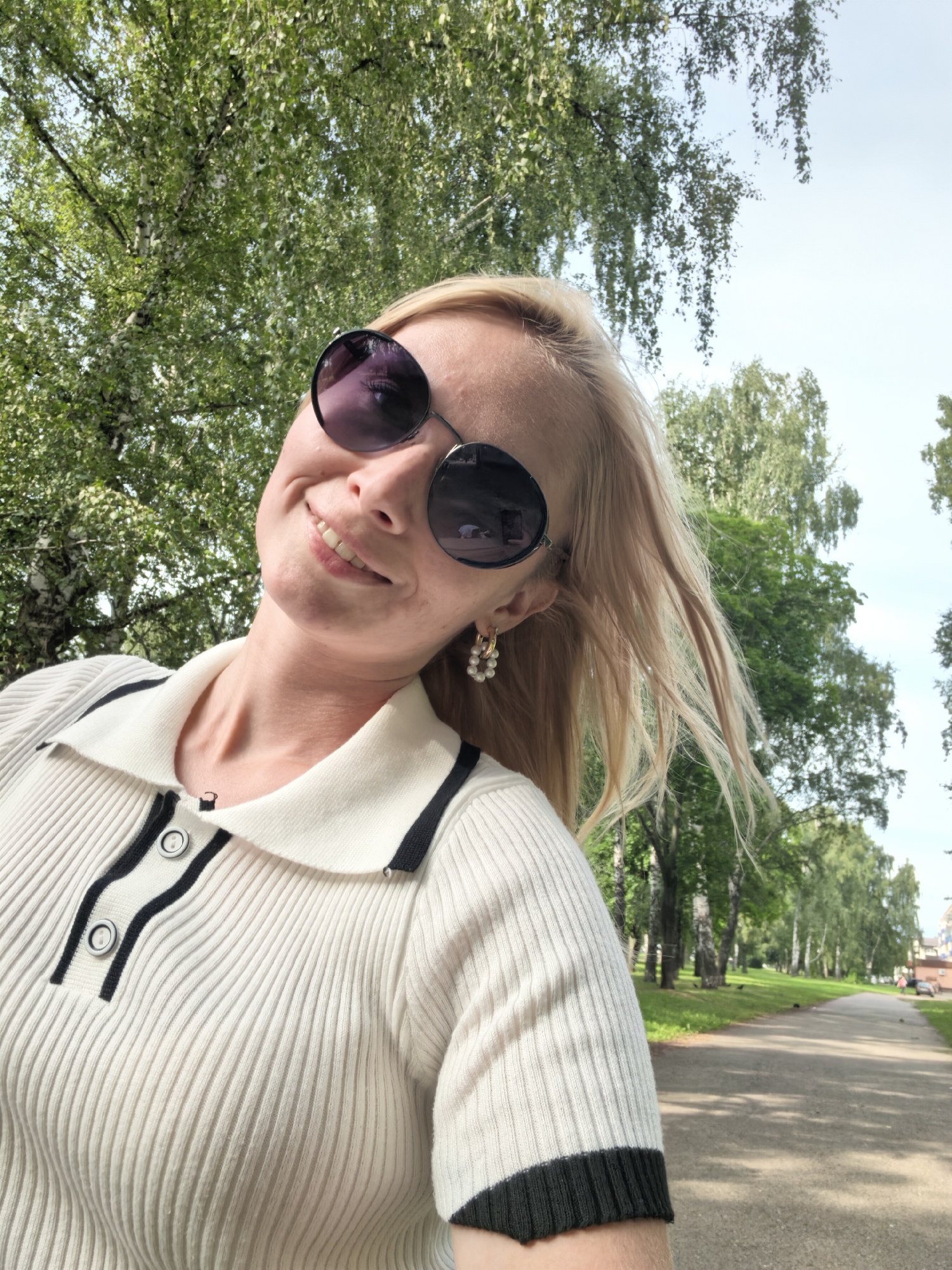 A medium shot captures a blonde woman with fair skin, wearing large, round sunglasses with purple lenses, and a white ribbed polo shirt with black trim and buttons. She is outdoors, with her head tilted back and a broad smile on her face. Her blonde hair is blowing gently in the wind. Behind her, a tree-lined path stretches into the distance, with a patch of green grass to the left and a clear sky above. The scene appears to be a park or a tree-lined street in a city. No specific landmarks or details are visible to pinpoint the location within Sterlitamak, Russia.