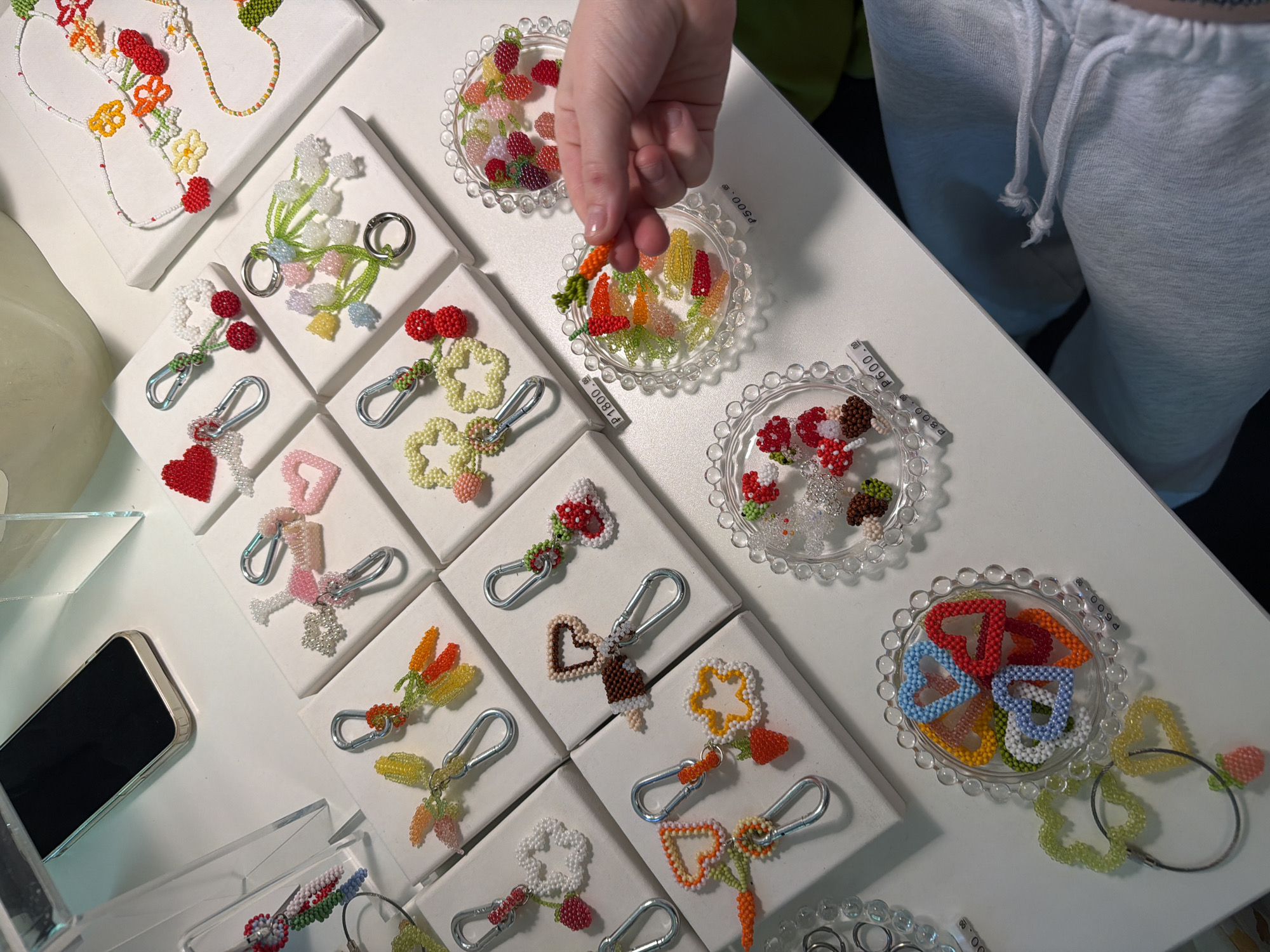 A high-angle shot captures a display of handmade beaded keychains and necklaces on a white surface. A person's hand is selecting a beaded carrot keychain from a clear, scalloped-edged dish. The keychains are displayed on small white square stands. To the left, a black smartphone sits on the surface. A person wearing light gray sweatpants stands to the upper right. Price tags are visible near some of the beaded items.
