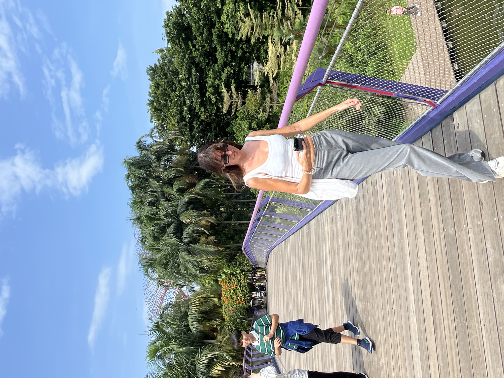 This is a medium shot of people walking across a wooden bridge with purple railings in Gardens by the Bay, Singapore. The woman in the foreground is wearing a white tank top, grey pants, sunglasses, and white sneakers. She is holding a phone in her right hand and a white tote bag is slung over her left shoulder. To her left, a young boy in a green and white striped polo shirt and black shorts walks towards the camera, with a blue jacket tied around his waist. Further back, a group of people are visible walking in the distance. The bridge overlooks a body of water and lush greenery, with palm trees and other tropical plants forming the backdrop. The sky is blue with scattered clouds.