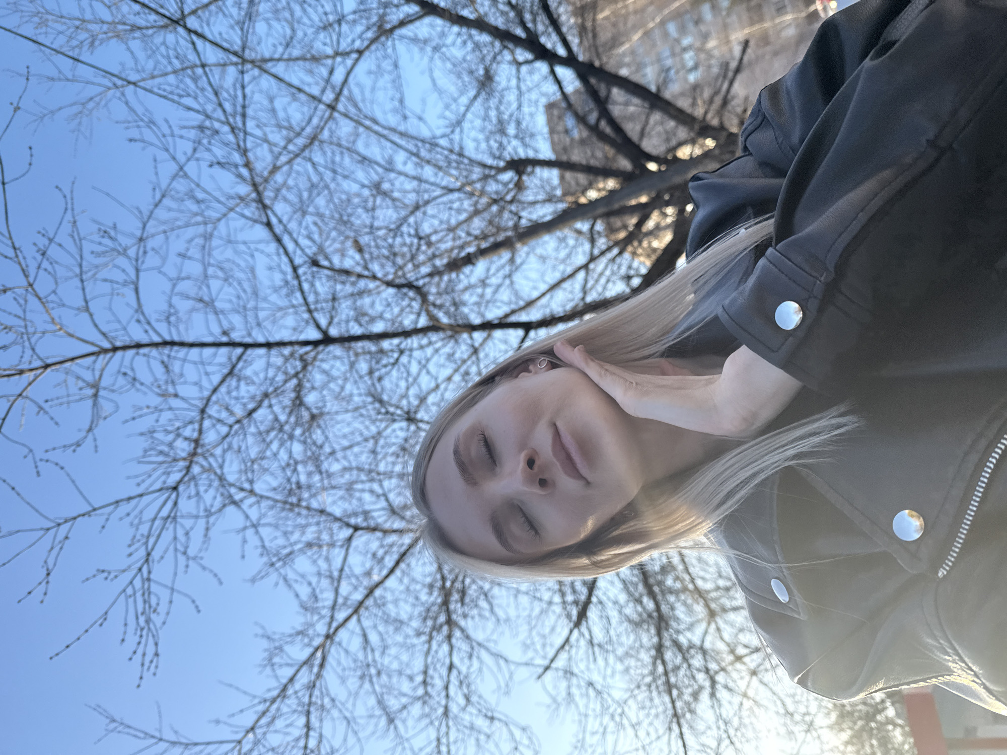 A young woman with blonde hair stands in what appears to be a park setting on a sunny day. She is wearing a black leather jacket with silver buttons and has her eyes closed, touching her face with her right hand. In the background, there are bare tree branches against a light blue sky. A tall building can be seen through the branches in the distance. The exact location within the city of Izhevsk, Russia cannot be determined from the image alone.