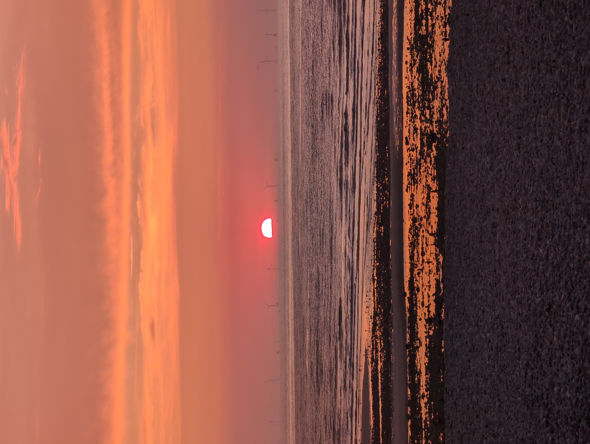 A vertical, low-angle, full shot captures a serene seascape at sunset in Le Tréport, France. The sky, painted in hues of orange and pink, is filled with horizontal streaks of clouds, creating a gentle gradient. The sun, a vibrant red orb, is partially submerged on the horizon, casting a warm glow. In the distance, a line of wind turbines stands silhouetted against the fading light. The sea is calm, with gentle waves lapping the shore. The foreground is a pebbled beach, with tide pools reflecting the colorful sky and scattered rocks. A few small birds are visible near the water's edge.