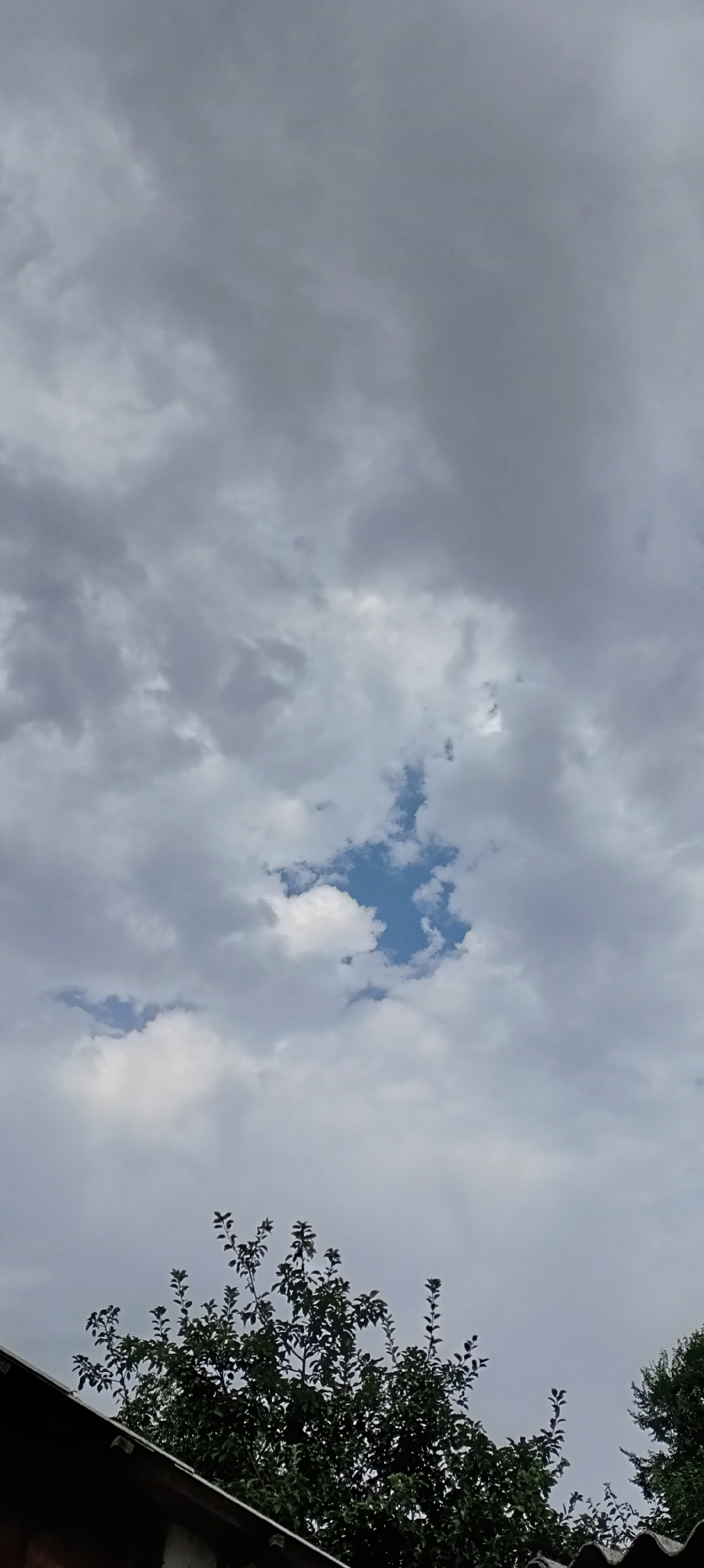 The image shows a view of the sky from the city of Sokuluk, Kyrgyzstan. A patch of blue sky is visible through a gap in the overcast, grey clouds. In the foreground, the tops of trees and the edge of a roof are visible. The scene appears to be outdoors, possibly in a residential area. There are no people or animals visible in the image.