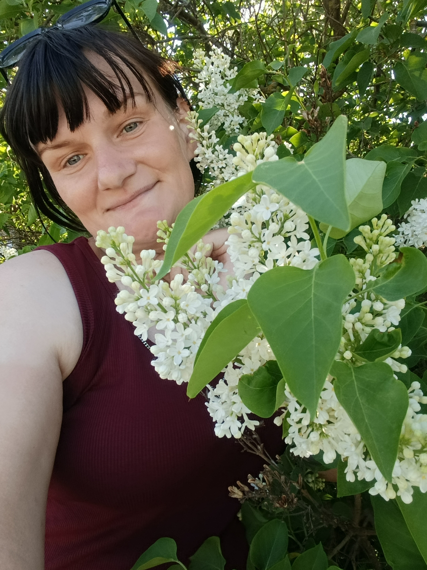 A woman with dark hair and bangs smiles into the camera while standing in front of a lilac bush with white flowers. She is wearing a maroon tank top and has sunglasses perched on her head. Her location cannot be determined from the image.
