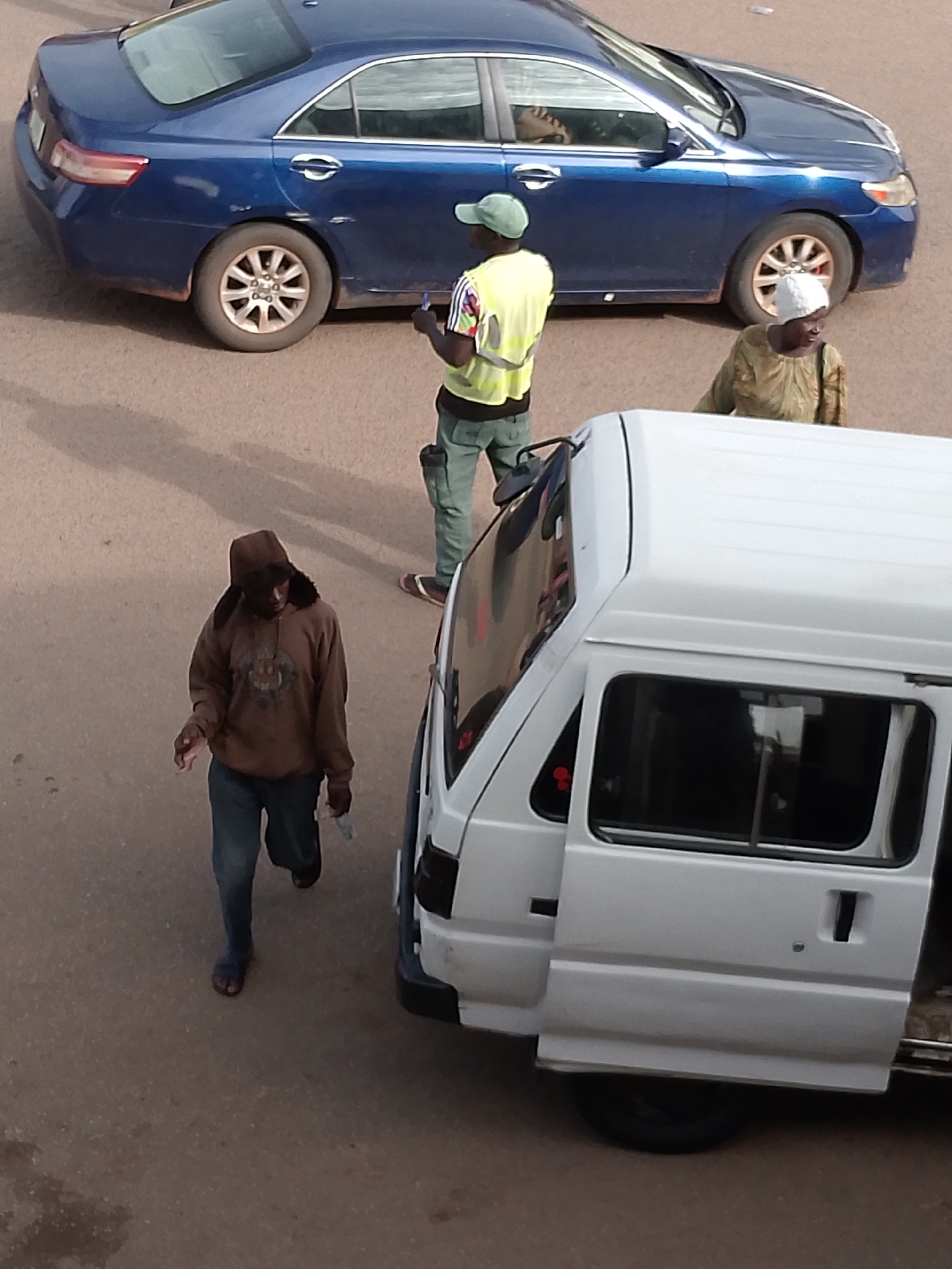 In Bukuru, Nigeria, a street scene unfolds with several individuals and vehicles. A dark blue sedan is parked on the asphalt road. To the right of the sedan, a person wearing a high-visibility yellow vest over a patterned shirt and green pants stands, holding an object. In the background, another person is partially visible, wearing a green garment and a white head covering. In the foreground, a white van with its side door open is positioned on the road. A person wearing a brown hooded sweatshirt, jeans, and sandals walks towards the left, holding a white object. The ground is a dusty, reddish-brown surface, with shadows indicating a bright light source.