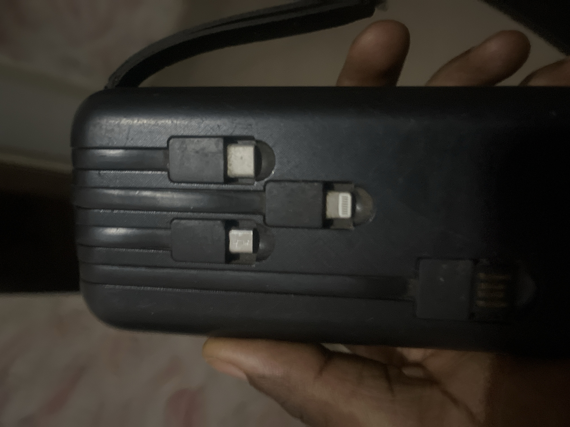 A close-up, eye-level, vertical shot shows a dark-colored, portable power bank held in the left hand of a person of color. The power bank has several integrated charging cables. Two cables with USB-C connectors and one with a Lightning connector are visible. A larger connector that appears to be a USB-A male end is also present. The background is blurred, with a pale, patterned surface on the left and a dark surface on the right. The scene is set indoors, likely in a residential or personal space within the city of Jalingo, Nigeria.