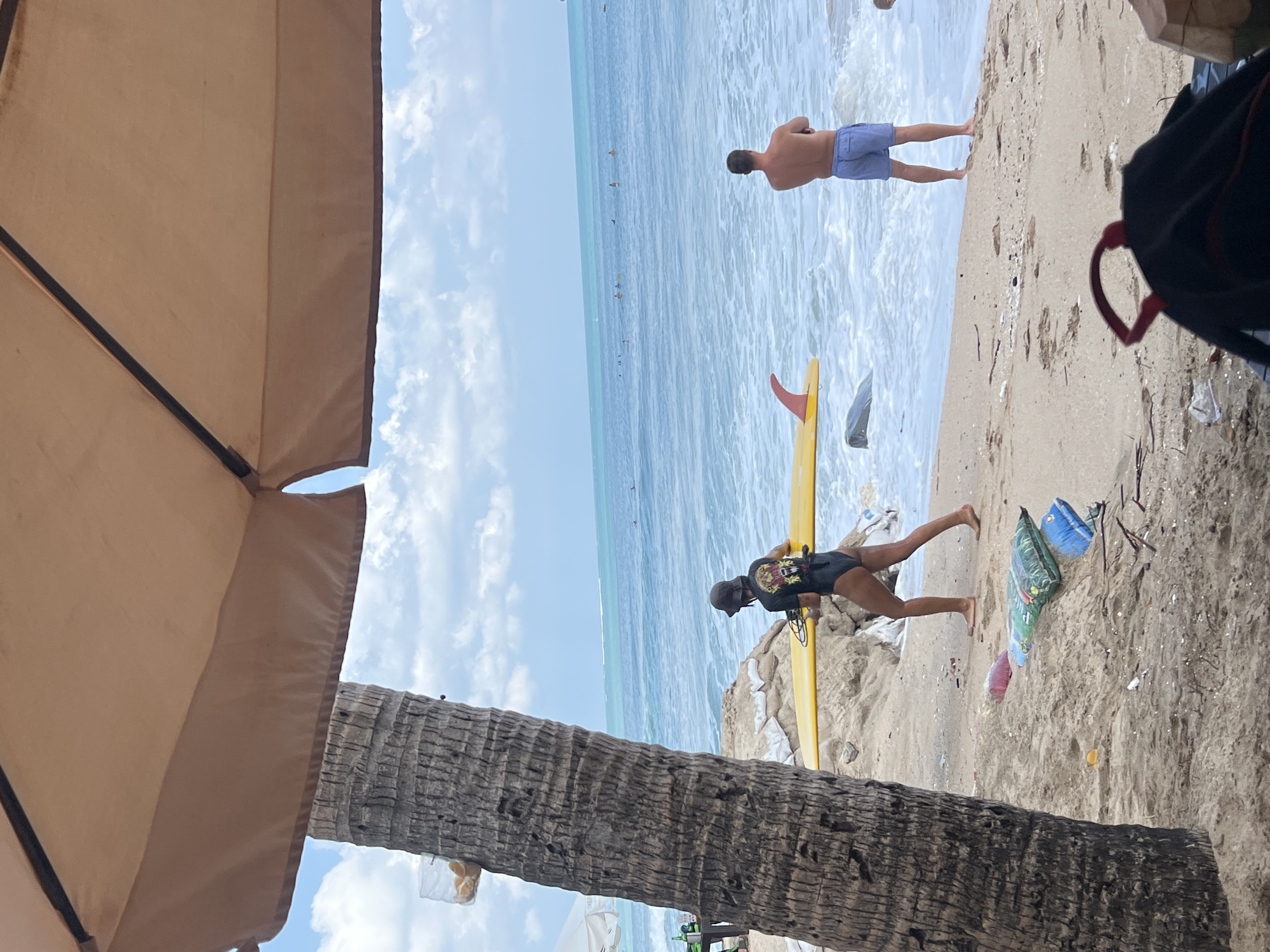 A beach scene in Kuta, Indonesia, unfolds under a large, light brown umbrella, partially obscuring the sky and a tall palm tree. The ocean, with gentle waves and a few surfers scattered in the distance, stretches to the horizon under a cloudy blue sky. On the sand, a woman wearing a black swimsuit and a bucket hat, carries a yellow surfboard. Nearby, a man in blue shorts stands facing the ocean, his arms crossed. Various objects litter the sandy shore, including sandbags, plastic bags, and debris. A coconut is visible in the lower right corner.