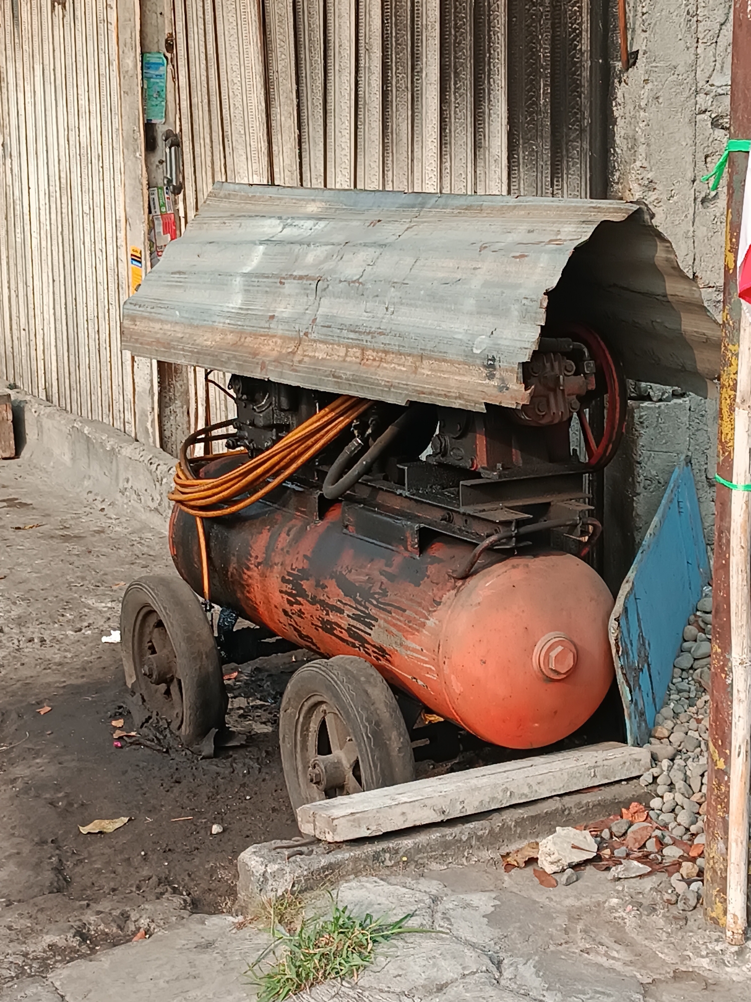 An orange and black industrial air compressor is parked outdoors in Banguntapan, Indonesia. The compressor, which has two wheels visible, is situated on a dirt and concrete surface. It is partially covered by a makeshift roof constructed from corrugated metal. The background consists of a grey concrete wall and a corrugated metal shutter. Scattered debris, including stones and dry leaves, is visible on the ground. The scene appears to be in an outdoor workshop or storage area.
