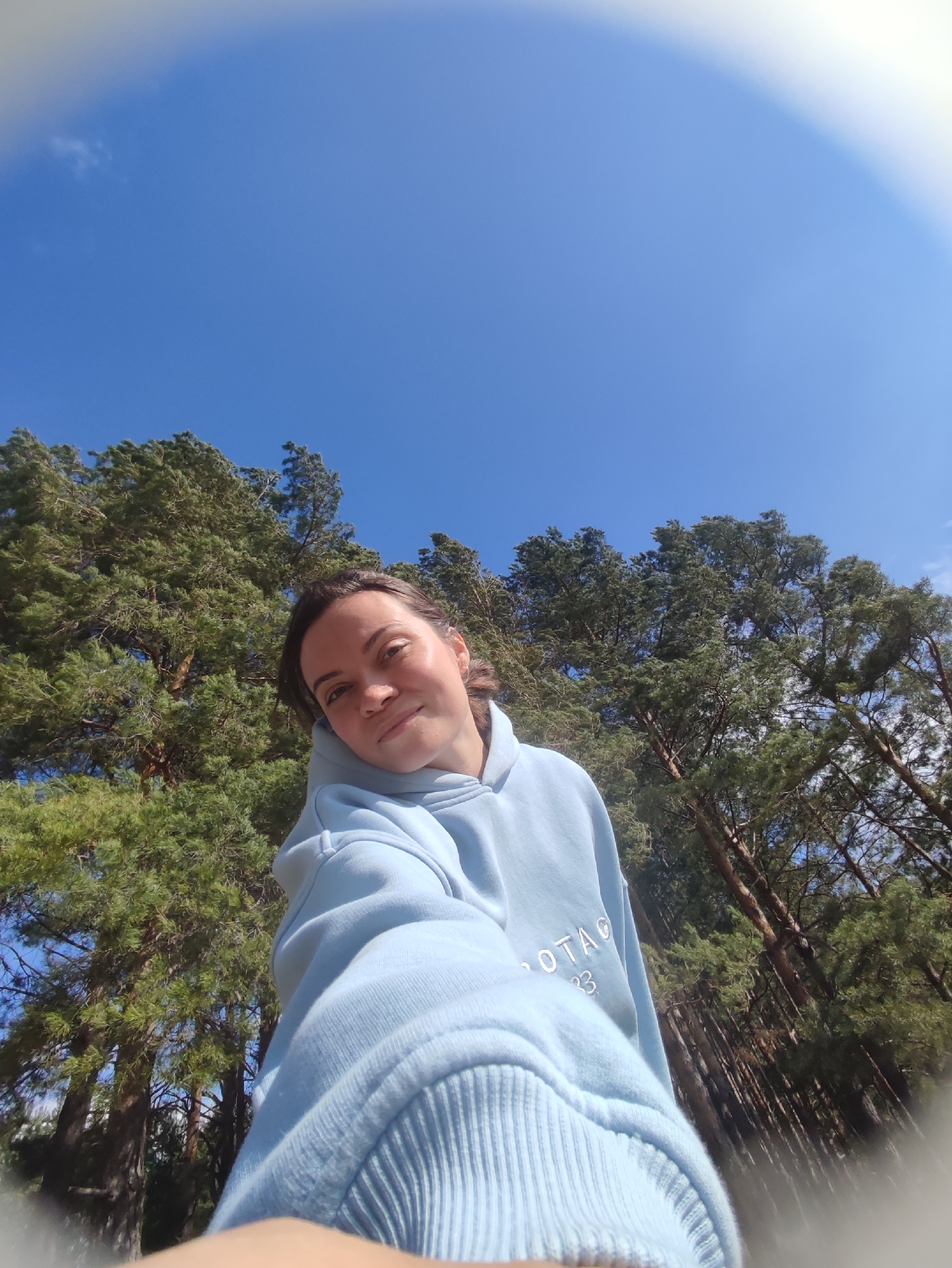 A selfie-style portrait captures a young woman outdoors on a bright, sunny day. The image is taken from a low angle, looking upwards towards a vast, clear blue sky with a few faint, wispy clouds visible in the upper left. The woman is the central subject, smiling gently at the camera, which she holds herself with an outstretched arm. She is wearing a light blue hooded sweatshirt, and on its front, partial text "OTAG" and "33" (or possibly "83") can be seen.
Behind her, numerous tall pine trees with dense green foliage dominate the background, suggesting a forest or wooded area. The bright sunlight illuminates her face and the tree canopy. The edges of the photograph display a slight circular distortion and blurring, indicative of a wide-angle or fisheye lens effect. The overall scene conveys a sense of peace and natural beauty, characteristic of a pleasant day spent outdoors, possibly in Фоки, Russia.