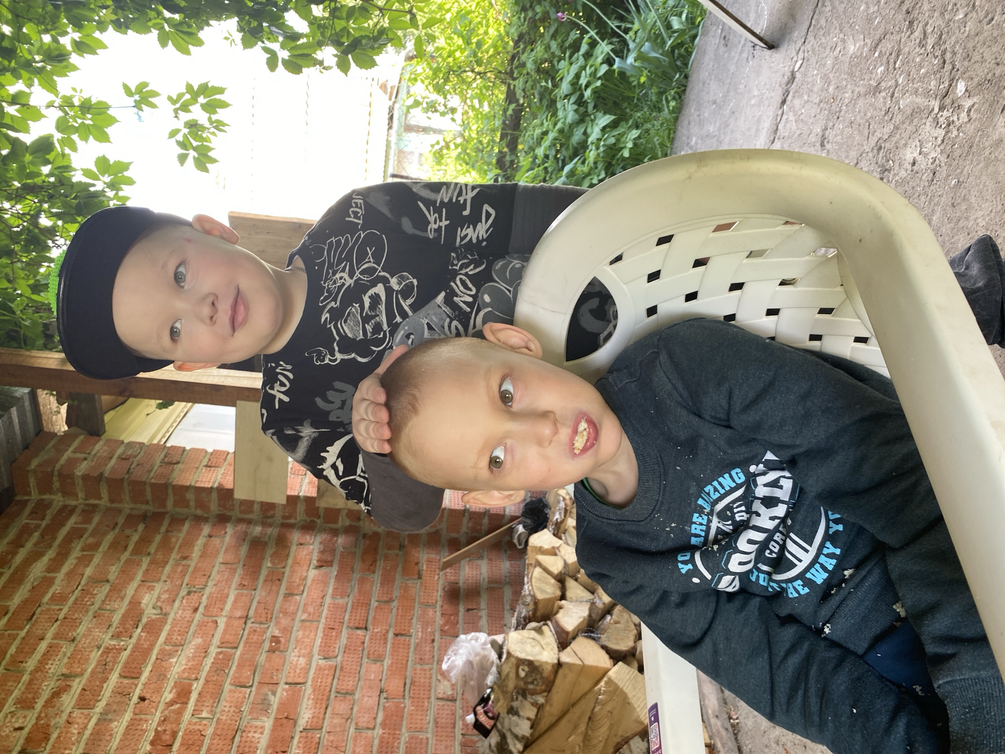 This image captures two young boys in an outdoor setting, likely a backyard or patio in Chaikovsky, Russia. The scene is brightly lit by natural daylight, suggesting a sunny or clear day.
The boy on the left, slightly behind, appears to be older. He wears a black cap and a black long-sleeved shirt with white graphic designs that include text like "FOR FUN" and partially visible letters "ECT". He has light-colored hair and eyes, and looks directly at the camera with a soft, friendly smile. His hands are positioned playfully near the head of the second boy.
The boy on the right, in the foreground, is younger and sits on a white plastic chair. He wears a dark grey long-sleeved sweatshirt with prominent blue and white text and graphics. Visible phrases on his sweatshirt include "YOU ARE AMAZING", "BROOKLYN", and "THE WAY YOU". There are small white particles, possibly crumbs, on his sweatshirt. He has a more energetic and playful expression, with wide, curious eyes and his mouth slightly open, revealing teeth with what appears to be food residue.
The background features a red brick wall on the left, and a neatly stacked pile of firewood logs on the ground towards the right. Lush green foliage and trees are visible overhead, indicating a natural outdoor environment. The ground beneath them is concrete. The overall mood of the image is candid and cheerful, capturing a lighthearted moment between the two boys.