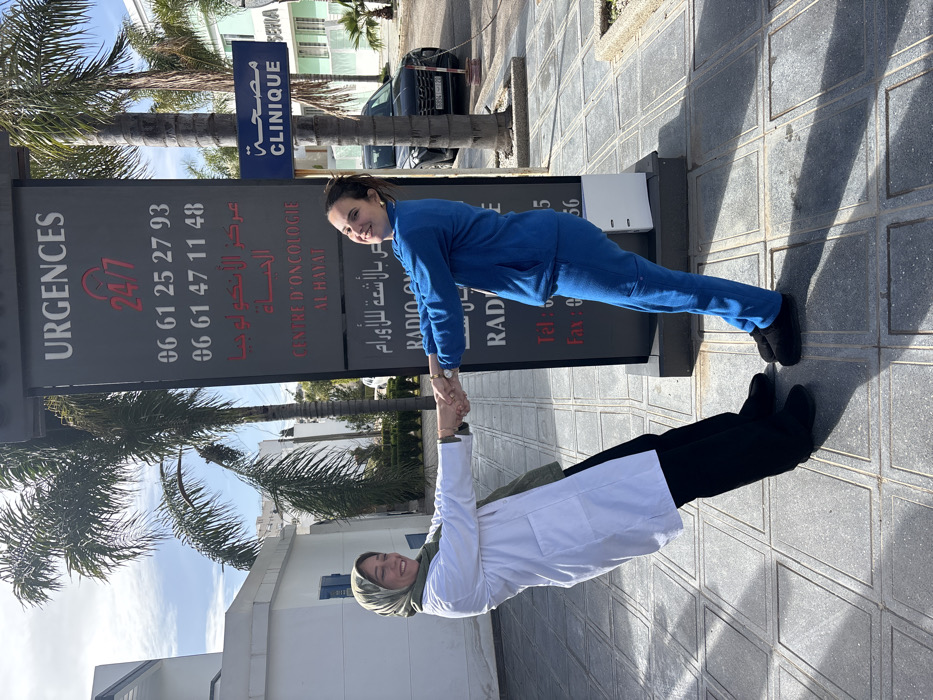 This outdoor scene depicts two adult females engaged in a playful interaction at the Centre d'Oncologie Al Hayat in Meknes, Morocco. The individual on the left wears a white lab coat over black pants and a green hijab, while the individual on the right is dressed in a blue scrub suit. Both subjects are smiling, leaning backward from a central vertical clinic sign, and holding each other's hands with outstretched arms, creating a symmetrical V-shape.
The prominent, dark grey vertical sign features white and red text, stating "URGENCES 24/7", followed by two phone numbers ("06 61 25 27 93" and "06 61 47 11 48"). Below this, it identifies "CENTRE D'ONCOLOGIE AL HAYAT" in French and Arabic. A smaller, blue rectangular sign positioned to the right of the main sign reads "CLINIQUE" in both French and Arabic.
The background includes a white building with a blue-shuttered window on the left, and a light green building on the right, both appearing to be part of the medical facility. Multiple palm trees are visible, with one positioned directly behind the main sign. A dark-colored SUV is parked on the street in the right background. The ground is paved with grey square tiles, exhibiting distinct shadows cast by bright sunlight under a partly cloudy sky.