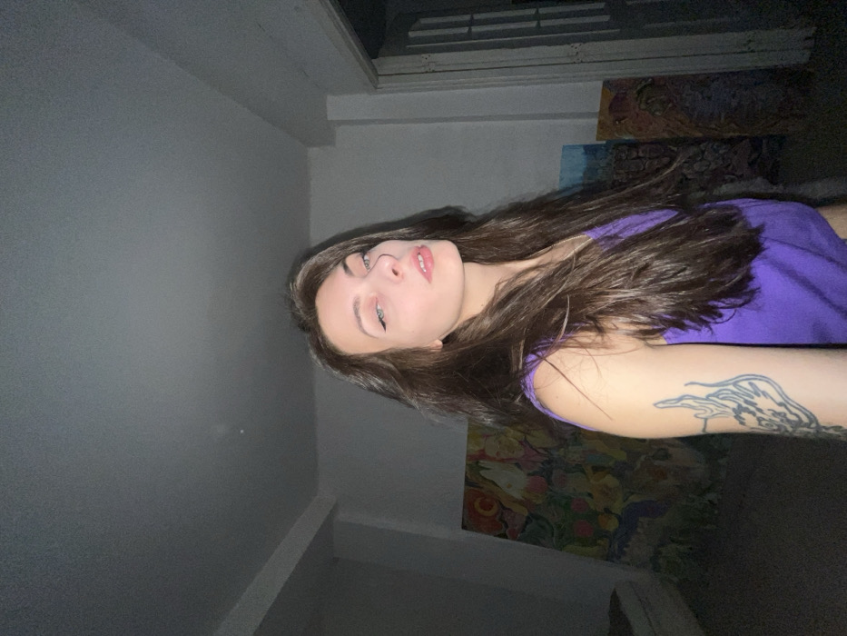 A woman with long brown hair and green eyes poses for a selfie. She's wearing a purple top, and a tattoo is visible on her arm. The background is dimly lit, showing a wall and window. There appear to be paintings or artwork hanging on the wall behind her. The angle of the photo is slightly tilted.