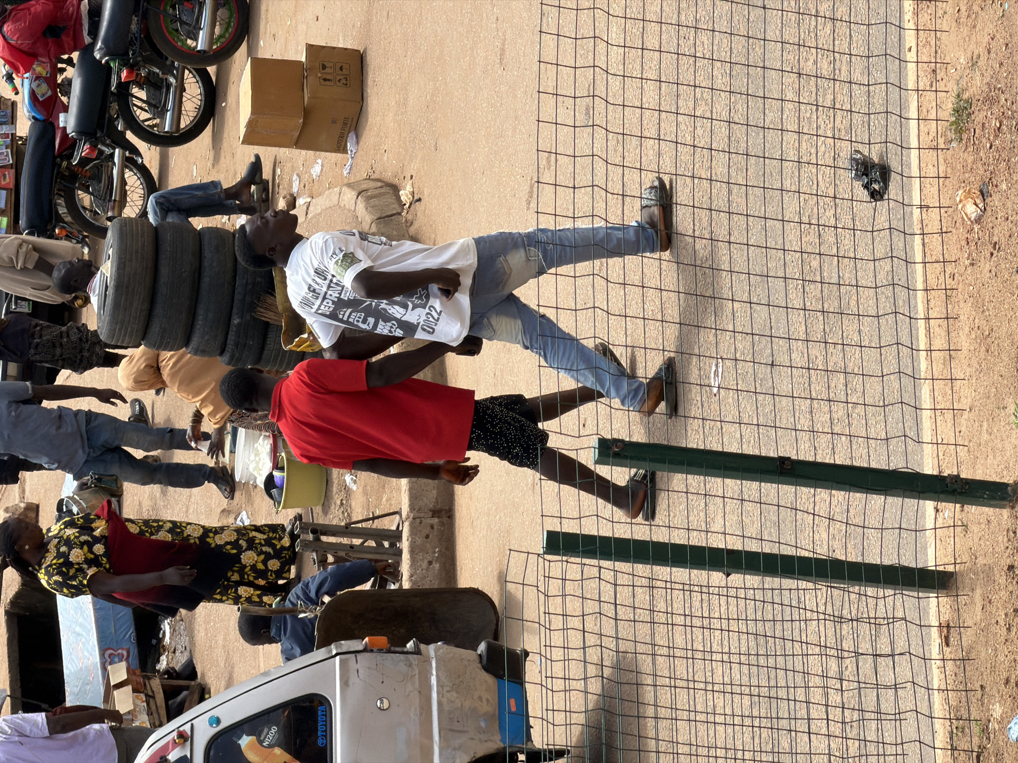 In Bukuru, Nigeria, this outdoor scene captures a bustling street with various individuals and objects. In the foreground, a metal fence with green posts is prominent. Behind the fence, two young men are walking on a paved surface. The man on the left wears a red t-shirt and black shorts, while the man on the right is in a white t-shirt and blue jeans. They appear to be walking together.
Further back, several other people are visible. A woman in a floral dress stands near a stall with a mattress. A man in a blue shirt and jeans is seated. There are stacks of tires and several motorcycles in the background, suggesting a marketplace or transport hub. Cardboard boxes are stacked on the ground near some of the motorcycles. The environment appears to be dusty with patches of sand and sparse vegetation. The overall impression is of a busy street scene in an urban setting.
