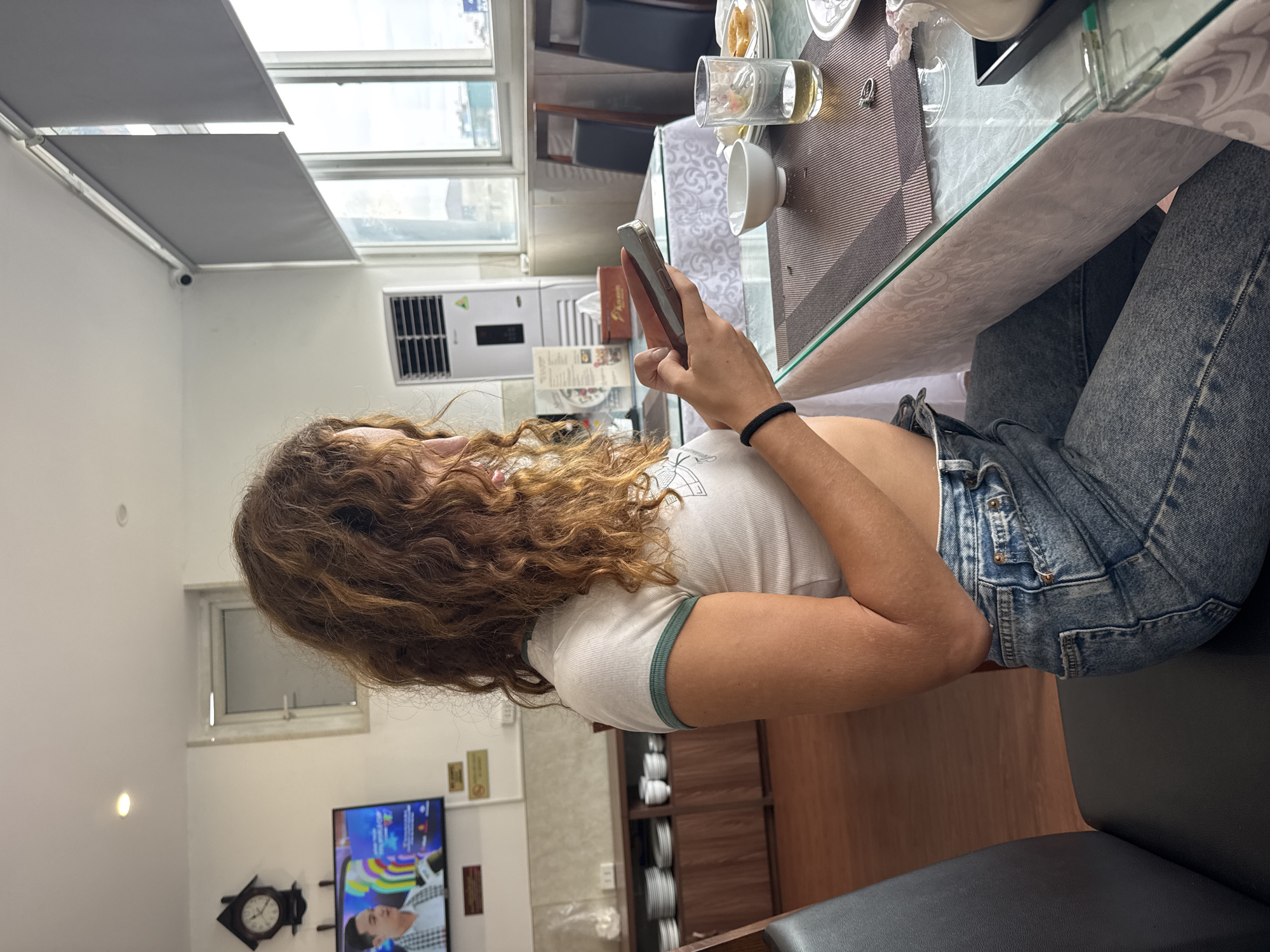 A young woman with curly brown hair, wearing a white crop top with green trim and blue jeans, sits at a table in a restaurant, looking at her phone. She has a black hair tie on her wrist. The table is set with placemats, bowls, plates, and glasses. In the background, there is a wooden cabinet with stacked plates and bowls, a television displaying a man's face, a clock, and a window with a view of buildings. An air conditioning unit is visible on the wall. The environment suggests a casual dining establishment.