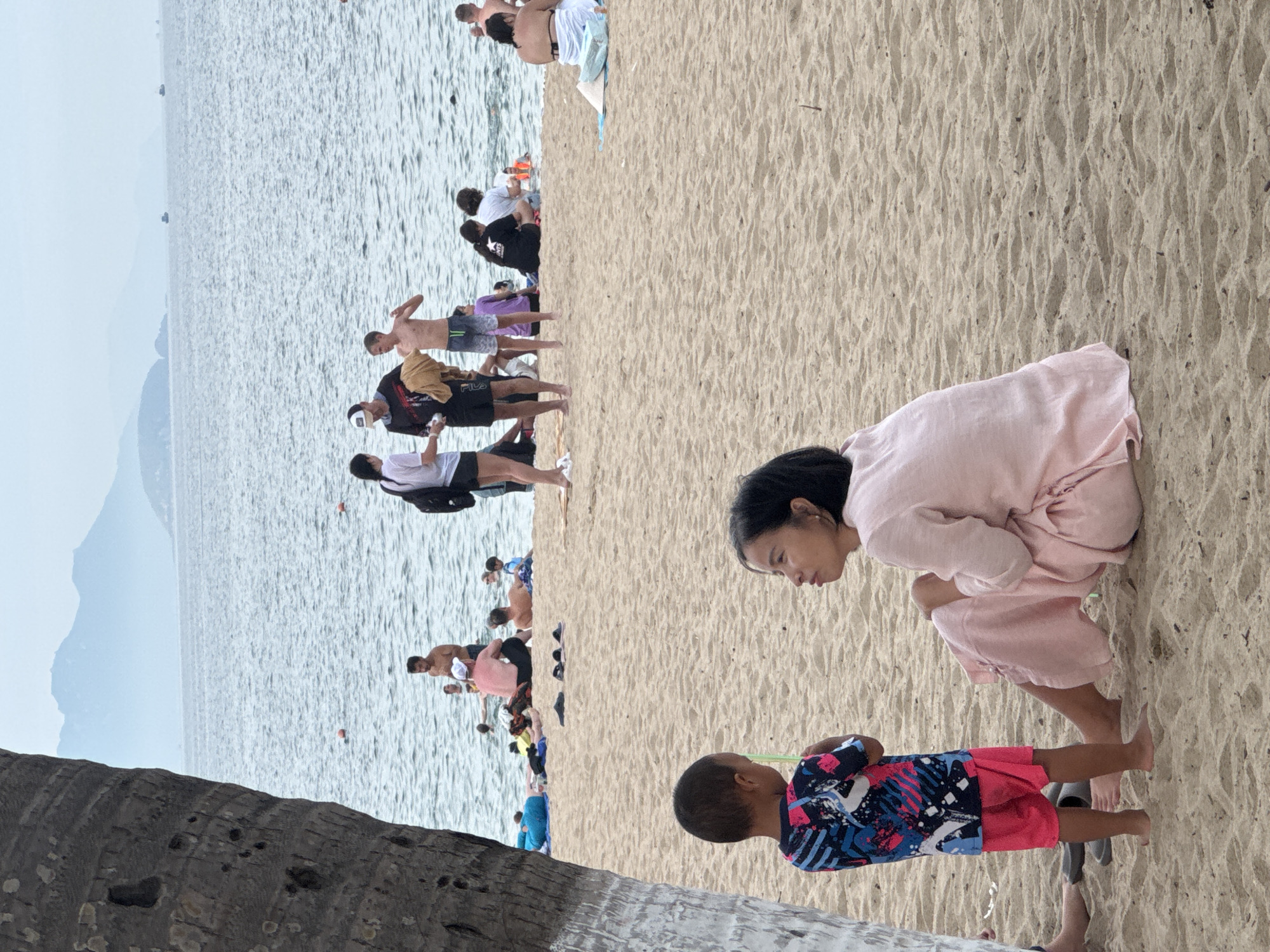 The image depicts a crowded beach scene on a cloudy day in Nha Trang, Vietnam. In the foreground, a young child wearing a brightly colored shirt and pink shorts stands facing away from the camera, holding a green straw. To the right, a woman in a pink dress sits on the sand, looking towards the child. The beach is populated with numerous people, some of whom are in the water or sitting on towels. The ocean stretches into the background, with distant islands visible on the horizon. The sand is light brown and textured, with footprints and debris scattered across its surface. The overall atmosphere is one of leisure and recreation.