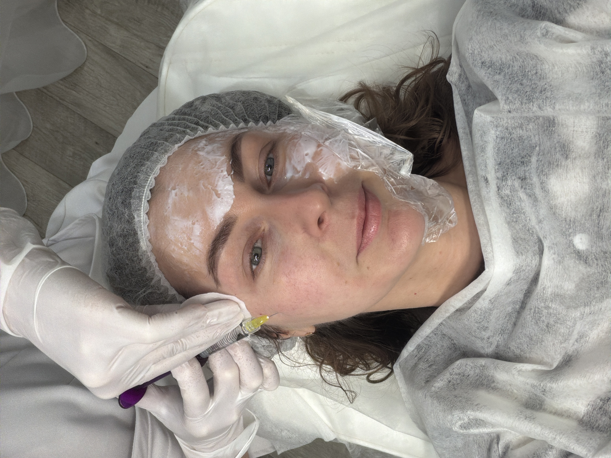 A woman wearing a disposable hair net and protective film over a white cream on her face is lying down, likely on a medical table covered with a white disposable sheet. A hand wearing a white glove is administering an injection with a syringe near her right cheek. The location is potentially a medical or cosmetic clinic in the city of Chaikovsky, Russia. The flooring is light grey wood-like planks.