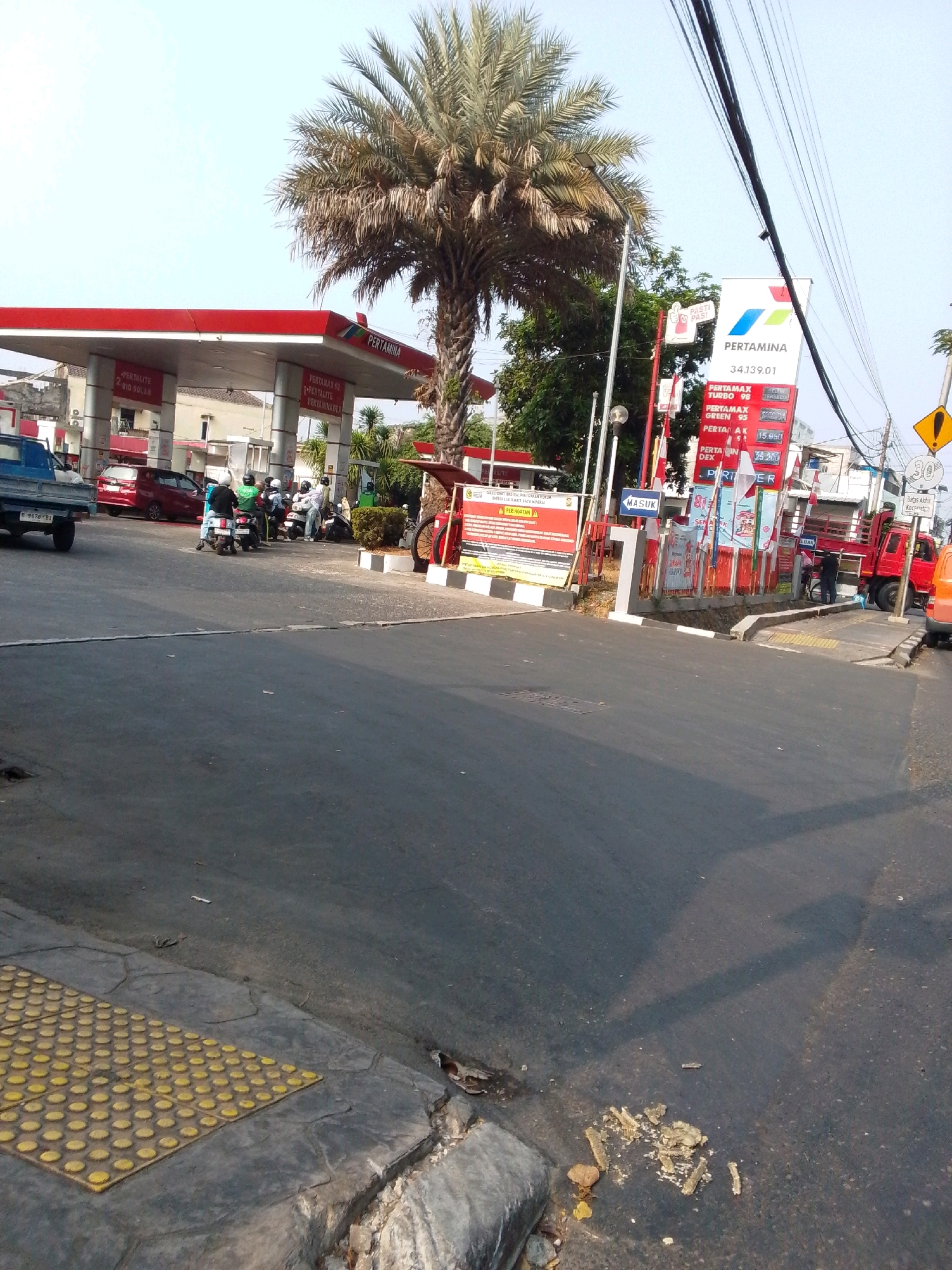 This is a medium shot, eye-level, outdoor, daytime, eye-level photograph taken at a Pertamina gas station in East Jakarta, Indonesia. The scene depicts a typical street view with a gas station on the left and a main road in the foreground and middle ground.
In the foreground, the asphalt road is visible, with a yellow tactile paving section on the sidewalk on the left. A cracked concrete curb separates the road from the sidewalk. Trash is scattered on the road and sidewalk.
The middle ground shows the gas station with its red and white canopy. Several cars and motorcycles are present, with people attending to them. A blue pickup truck is parked on the left. On the right, a red truck is partially visible. A tall palm tree stands in front of the gas station canopy.
In the background, buildings and more trees are visible. The sky is clear and bright blue. A Pertamina price board is prominently displayed on the right, showing fuel prices. Traffic signs are also visible, including a speed limit sign of 30.
The overall impression is a busy, everyday scene at a gas station in an urban environment.