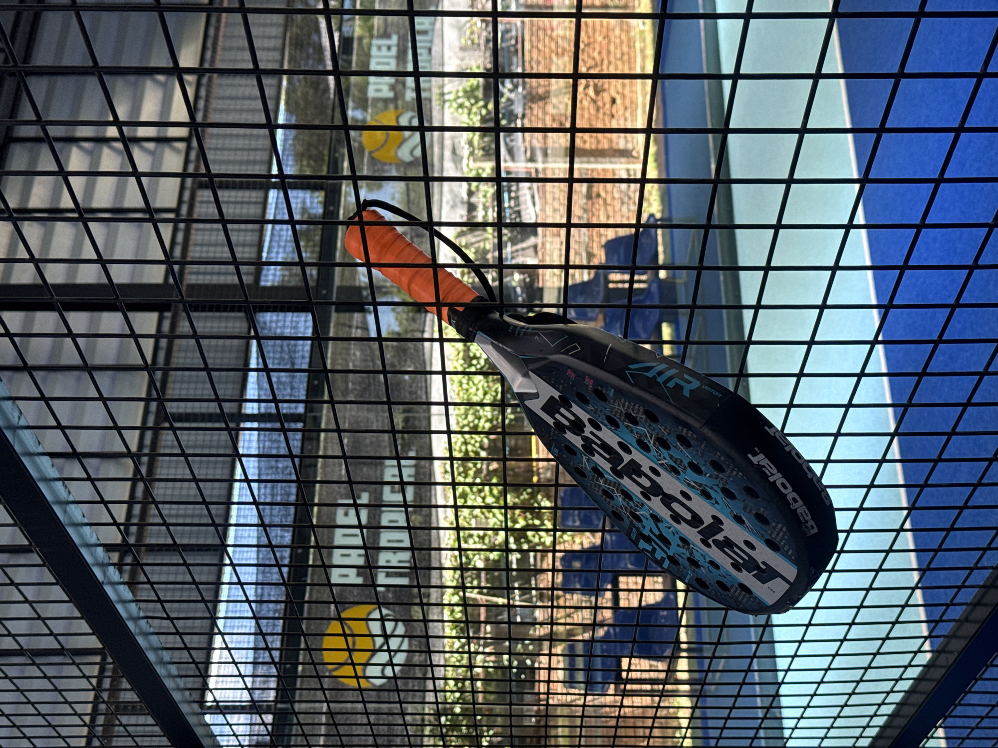 A padel racket, predominantly black with turquoise accents and white lettering that reads "Babolat," hangs from a black wire fence. The racket has an orange grip and is angled downwards, with its head slightly obscured by the fence. Behind the fence, a blue padel court is visible, partially illuminated by sunlight. In the background, there are rows of blue chairs and a brick wall. A sign with a yellow and white logo and the words "PADEL TROPICAL" is visible on the fence to the left. The scene is outdoors, likely at a padel club in Ban Bang Rak, Thailand, given the context provided.