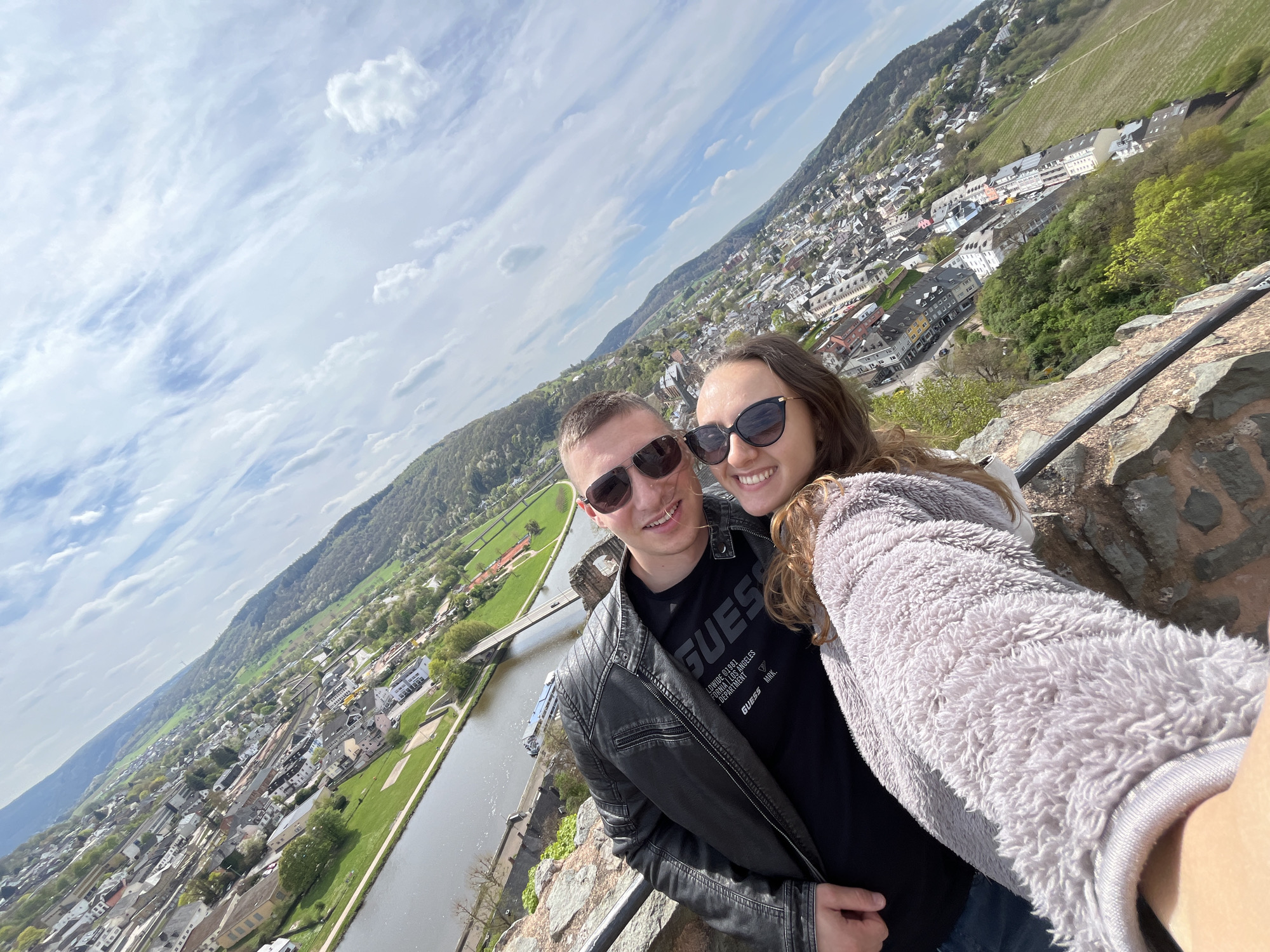 A selfie of a smiling man and woman wearing sunglasses. The man is in a black leather jacket and a black "Guess" T-shirt, and the woman is in a soft gray jacket. They are standing on a stone ledge or wall. Below them is a panoramic view of a European city alongside a river. A bridge is visible over the river. Hills covered with buildings and trees form the backdrop, with a sky with clouds. The town is identified as Saarburg, Germany.