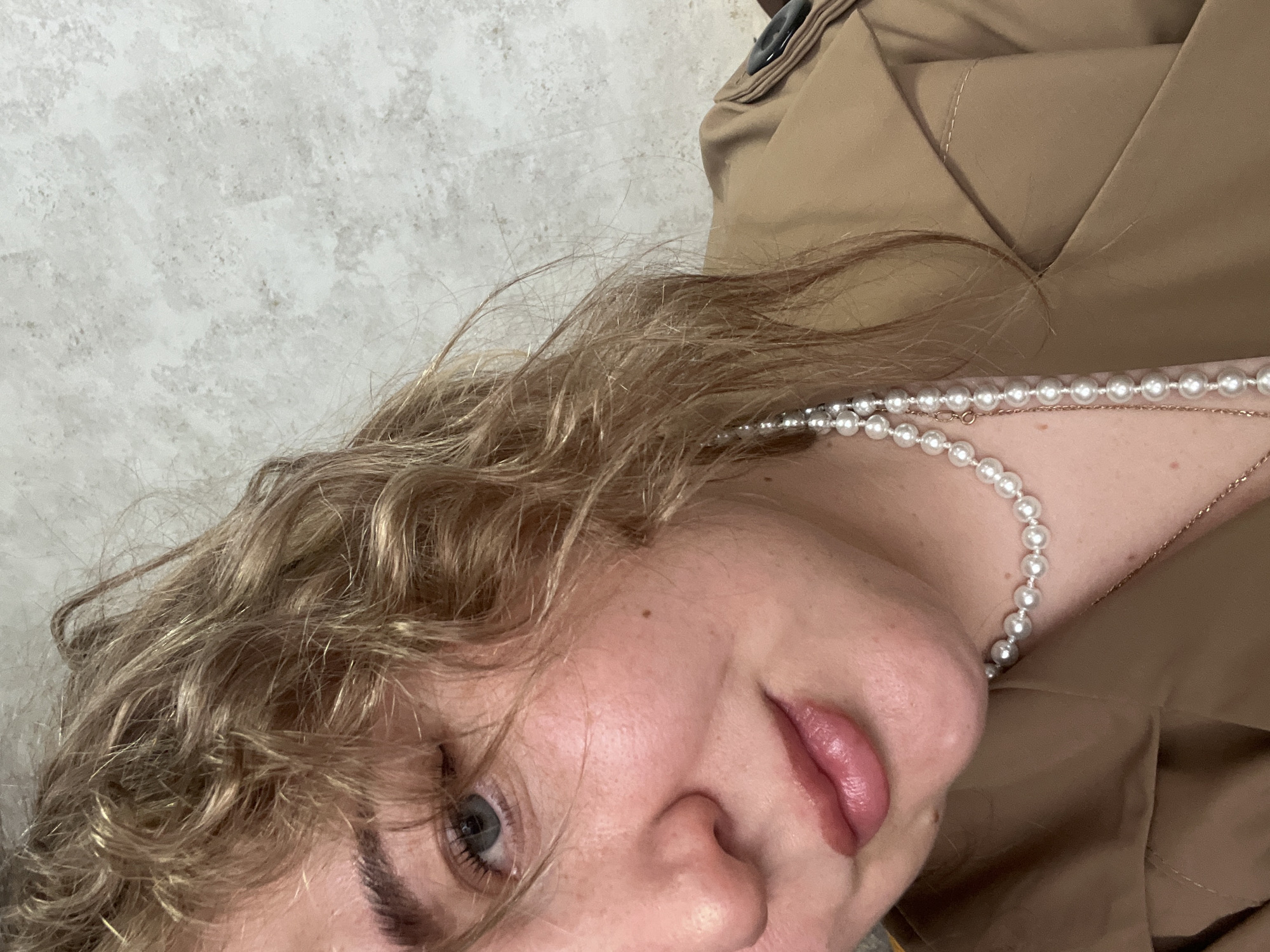 This is a close-up portrait of a person, likely a woman, with light brown, curly hair that frames her face. She has fair skin, light-colored eyes (appearing blue or grey), and full, slightly parted lips. A small mole or freckle is visible on her right cheek.
She is wearing a multi-strand pearl necklace and a delicate gold chain, both resting on her neck and collarbone. Her attire includes a light brown or tan-colored garment, which appears to be a coat or jacket, identifiable by its structured fabric, visible stitching, and a dark button near the shoulder.
The person is positioned diagonally within the frame, with her head tilted, and her gaze directed towards the camera. Her expression is calm or neutral. The background is a light-colored, textured wall, suggesting an indoor setting. The image is brightly lit. There is no visible text. The location context for this image is Chaikovsky, Russia.