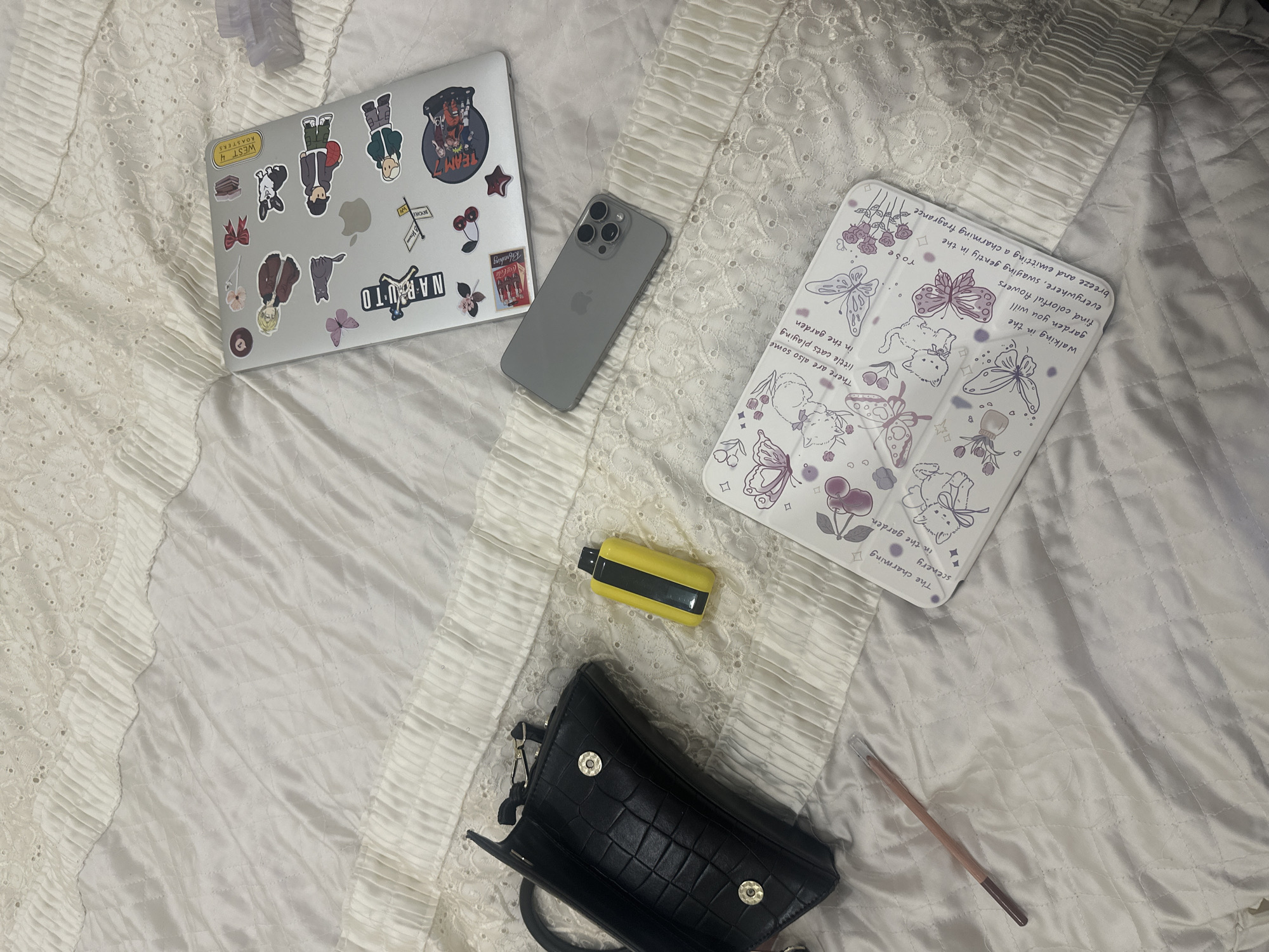 The image shows a cluttered bed with various items scattered on a white, textured bedspread. There's a laptop with stickers, a gray smartphone, a tablet in a patterned case, a yellow vape pen, a small black handbag, and a pink pencil. The laptop's stickers feature anime characters and a "WEST 4 ROASTERS" logo. The tablet case has a whimsical design with butterflies, flowers, and cats, accompanied by text describing a charming garden. No people or specific Yerevan city location details are discernible.