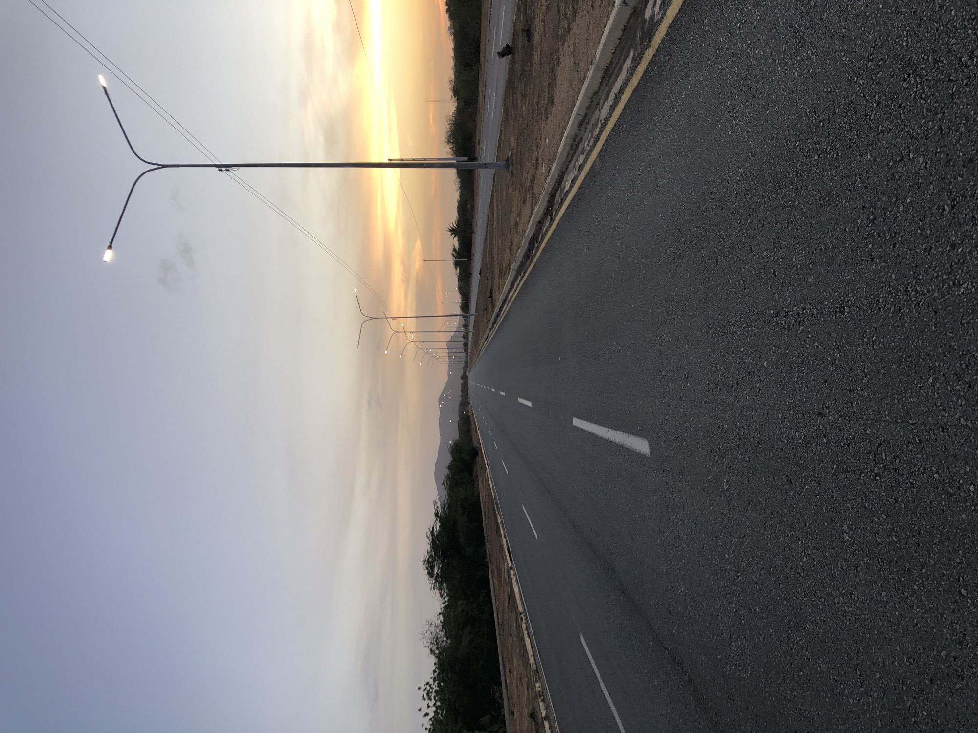 This image depicts a straight, paved road receding into the distance, lined with evenly spaced streetlights. The road has white lane markings and is bordered by vegetation on one side and a curb and unpaved shoulder on the other. The sky transitions from a light blue to a golden-yellow hue near the horizon, suggesting either sunrise or sunset. The setting could possibly be in the city of Altagracia, Venezuela, due to the presence of mountainous terrain in the distance. There are no visible people or active vehicular traffic in the immediate foreground.