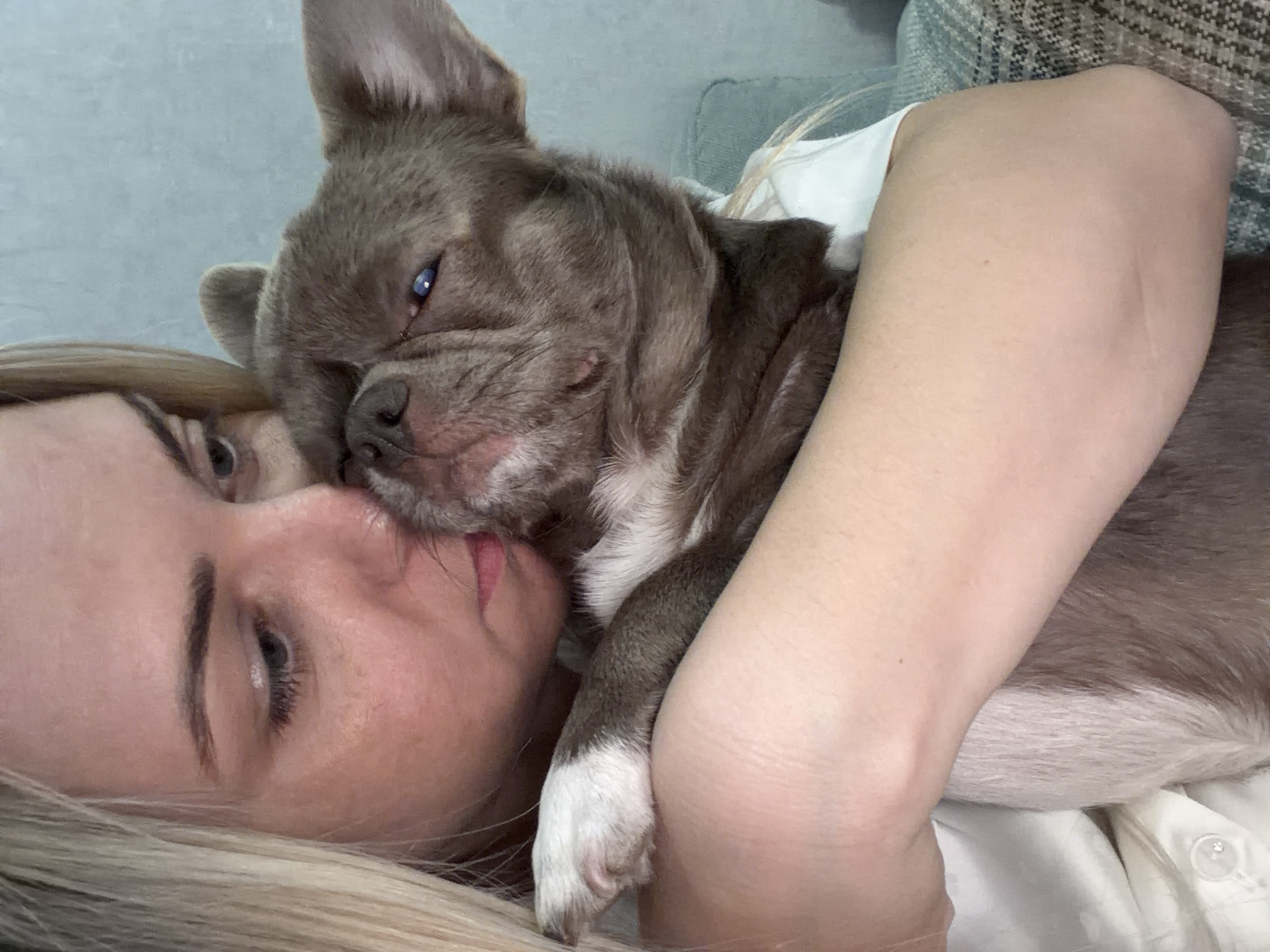 A close-up, high-angle image captures a tender moment between a person and a small, short-nosed dog, possibly a French Bulldog, in an indoor setting within Votkinsk, Russia. The person, appearing to be a woman with fair skin and long blonde hair, lies on their side, with their eye and defined eyebrow visible. Their lips are slightly parted, seemingly in a relaxed or affectionate pose.
The dog, with greyish-brown fur and distinctive white markings on its chest and paws, is snuggled intimately against the person's face and upper body. Its head rests near the person's mouth and nose, suggesting a nuzzle or close embrace. One of the dog's ears is partially visible, while its striking blue eye, reflecting light, is a notable feature. A white paw with dark pads is visible against the person's arm, which appears to be holding the dog.
The scene conveys a strong sense of comfort, affection, and peacefulness. The person is wearing a white garment, with a button visible on the sleeve. In the background, a soft, patterned fabric, possibly a couch or blanket, indicates a cozy, domestic environment. The lighting is soft, suggesting a relaxed time indoors. There is no visible text in the image.