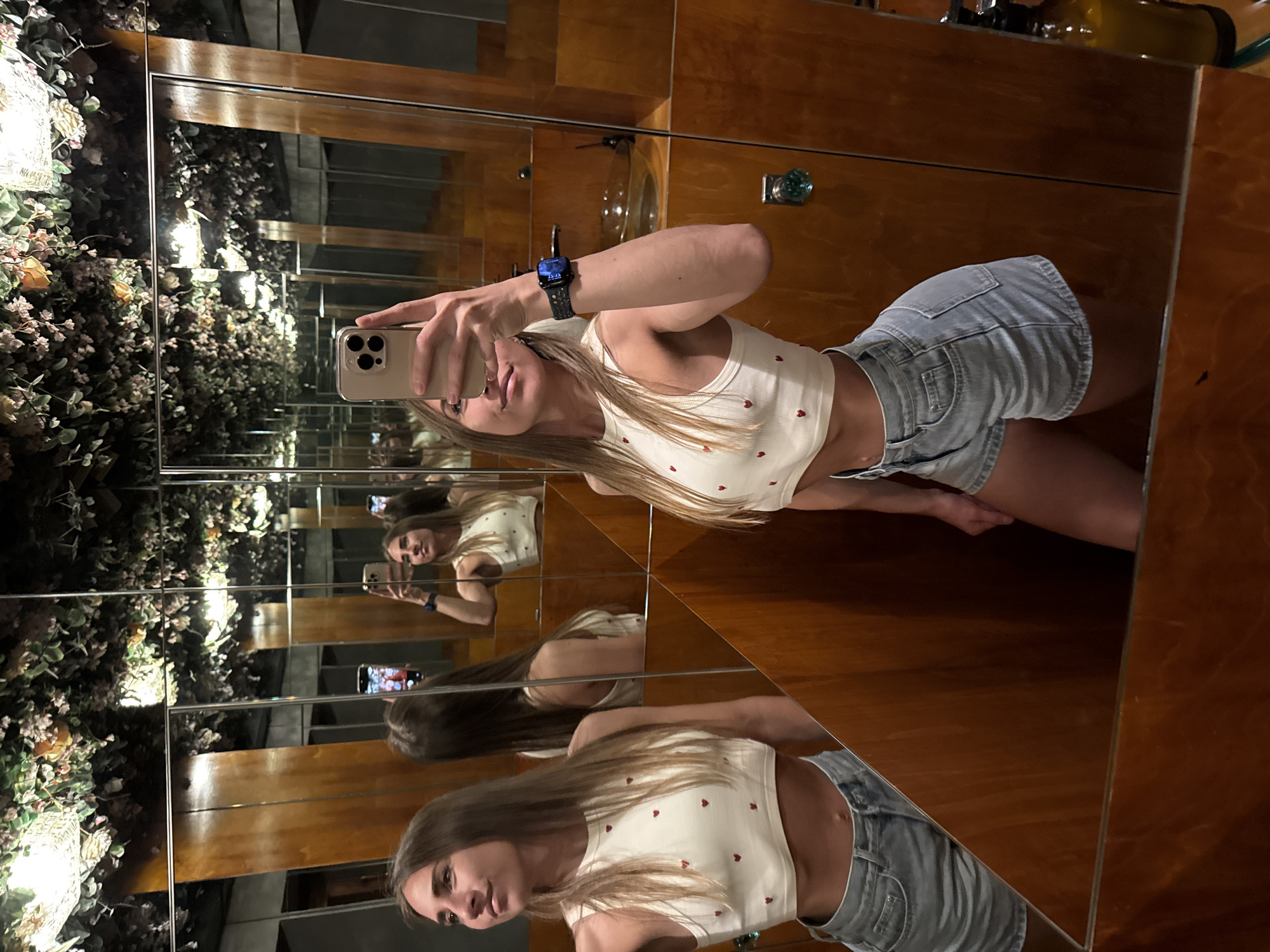 A woman is taking a selfie in a bathroom with mirrored walls. She is wearing a white crop top with red hearts and denim shorts. The bathroom features wooden cabinets and a ceiling covered in faux greenery and flowers. The woman is holding her phone up to take the photo, and her reflection is visible in multiple mirrors. The lighting is dim, and the overall atmosphere is intimate and somewhat surreal due to the reflections and the ceiling decor. The location is not specified as being in Saint Petersburg, Russia.