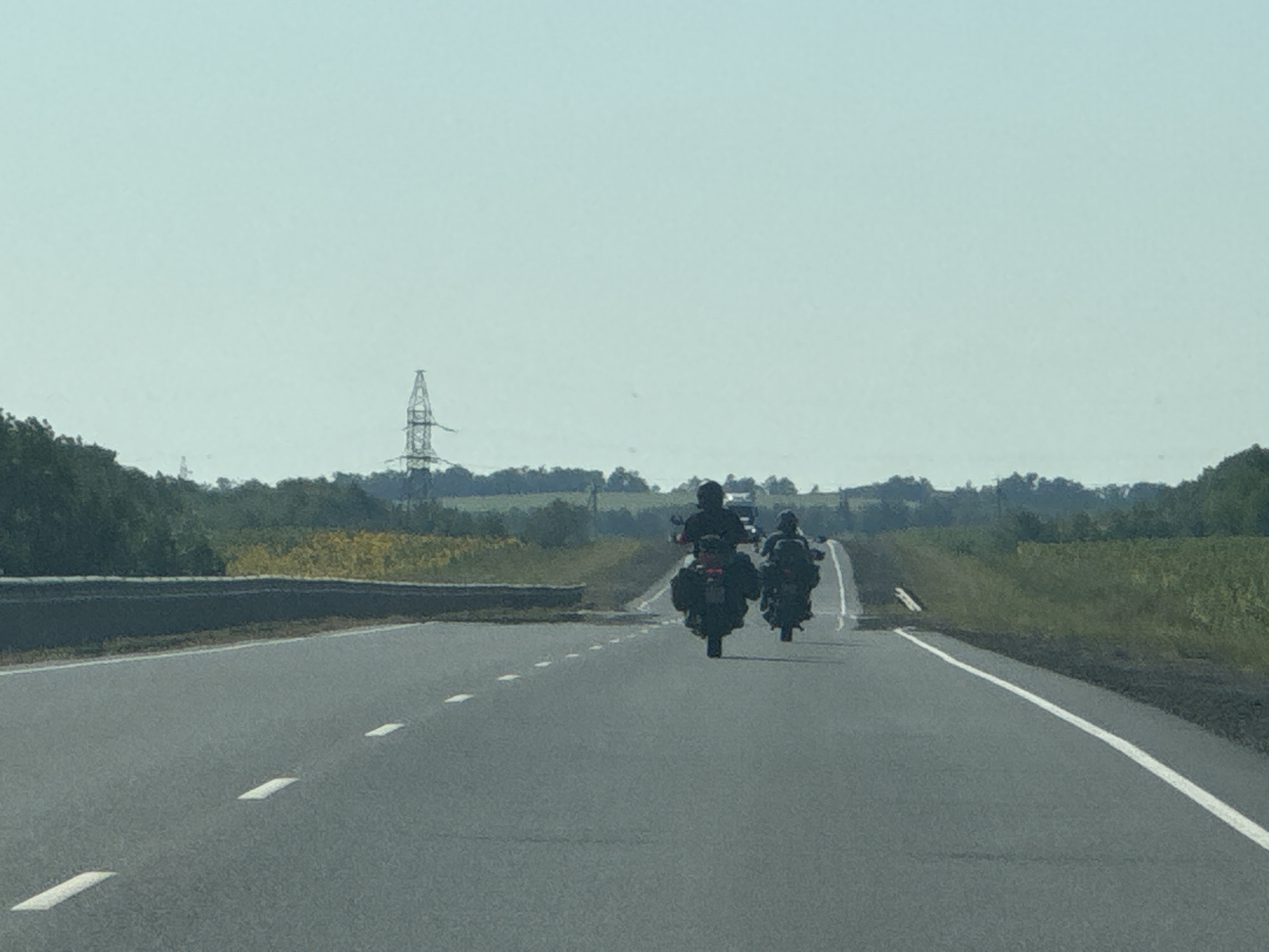 Two individuals are riding motorcycles on a paved road with white lane markings. The motorcyclists are wearing helmets and appear to be traveling away from the camera. The motorcycle in the foreground is closer and larger, with saddlebags. The second motorcycle is further in the distance. The environment is rural, with trees and fields visible on either side of the road. In the distance, a tall industrial structure, possibly a power line tower, is visible against a pale sky. The image was taken on a bright, clear day. The specific location in Perevolotsky, Russia, is not identifiable from the visual elements alone.