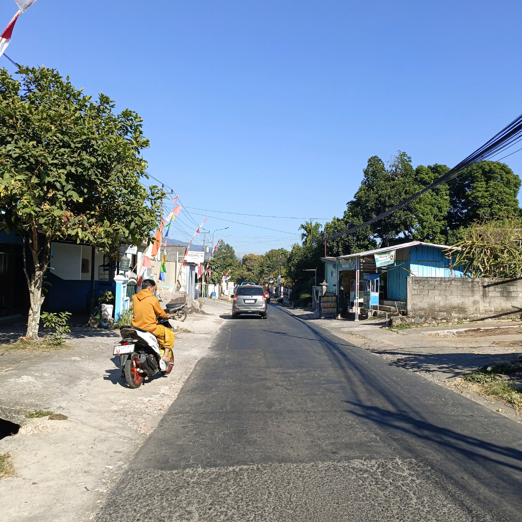 A sunny day is depicted in Sraten, Indonesia, with a clear blue sky dominating the upper portion of the image. In the foreground, a person is riding a white scooter on a paved road. The rider is wearing an orange hoodie and yellow pants. The road has patches of different textures, suggesting recent repairs. To the left of the road, there is a concrete sidewalk with a tree and some buildings. The buildings appear to be small shops or residences, some with blue accents. Small Indonesian flags are strung across the road in the middle ground, indicating a festive occasion or celebration. A silver car is driving on the road ahead of the scooter. The right side of the road features more buildings, including a light blue structure with a gray brick wall in front of it. Lush green trees are visible throughout the scene, and power lines stretch across the sky. The overall impression is a typical village street scene on a bright, warm day.