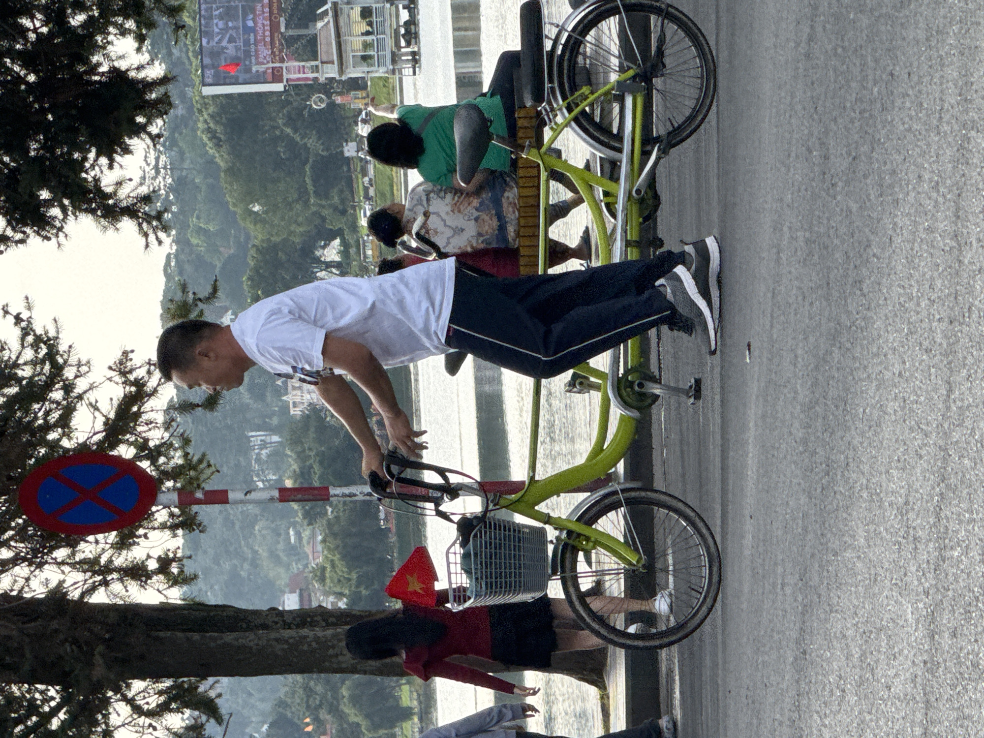 The image captures a scene on a street in Da Lat, Vietnam, likely near Xuan Huong Lake, characterized by a tree-lined path and a body of water in the background. A man in a white t-shirt and dark pants is riding a tandem bicycle, which is painted bright green. The bicycle has a basket on the front. Two women are seated on the back of the tandem bicycle. A woman in a red top and black skirt is walking away from the bicycle, holding a small Vietnamese flag. A stop sign with a red circle and blue cross is visible on a striped pole. In the background, there is a body of water, lush green hills, and some buildings. A large digital billboard displaying text and an image is also visible. The overall atmosphere appears to be a casual outdoor setting.